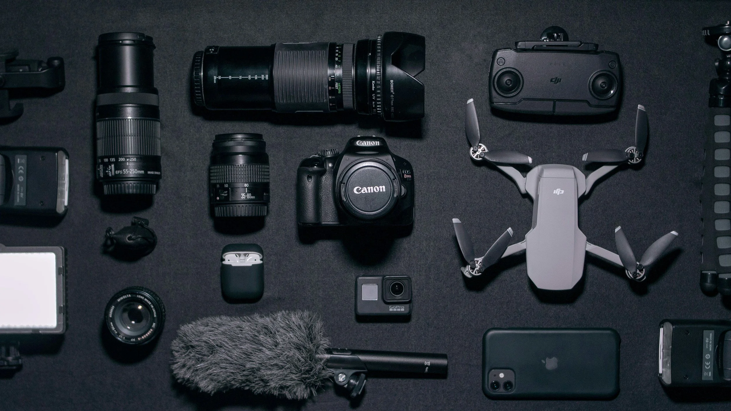 Collection of camera equipment including camera, lenses, drone, remote control, and accessories on a dark background.