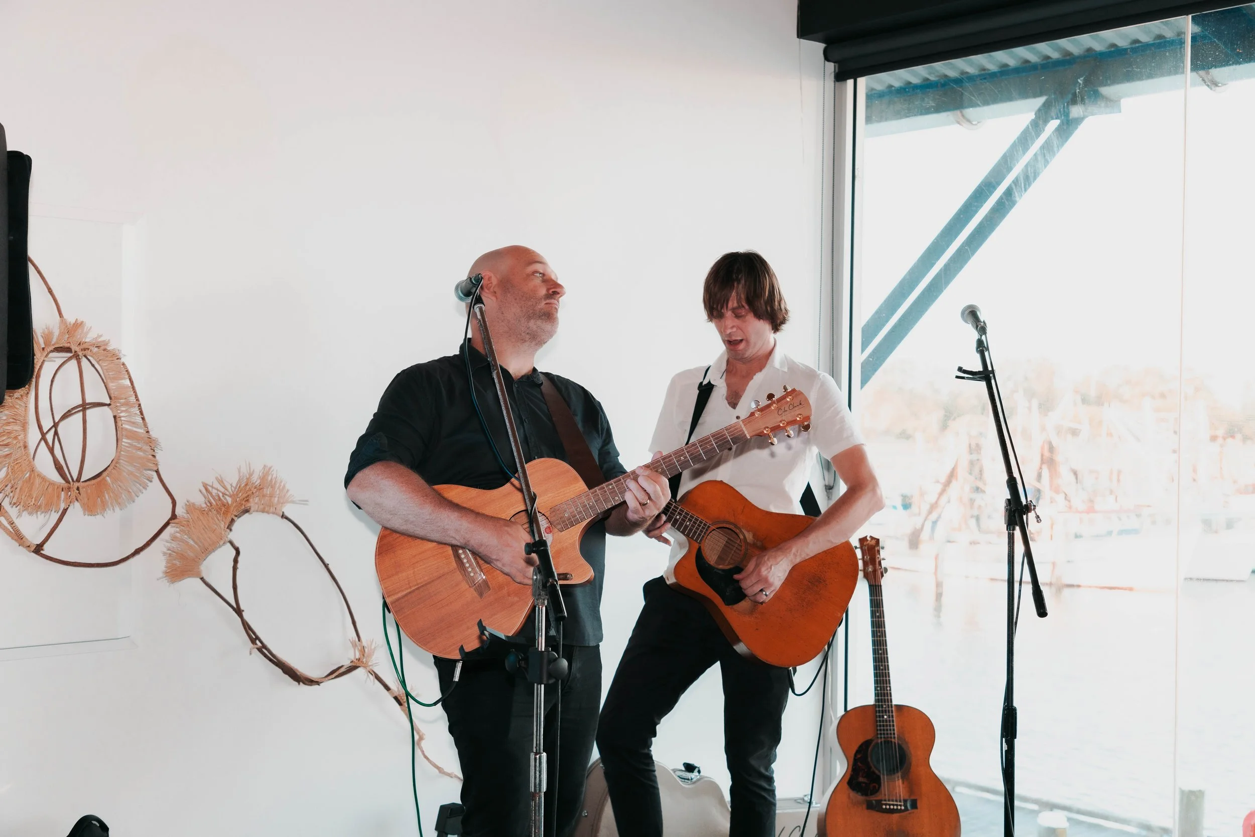 Two musicians playing acoustic guitars during a live performance indoors near a window with a view of boats.
