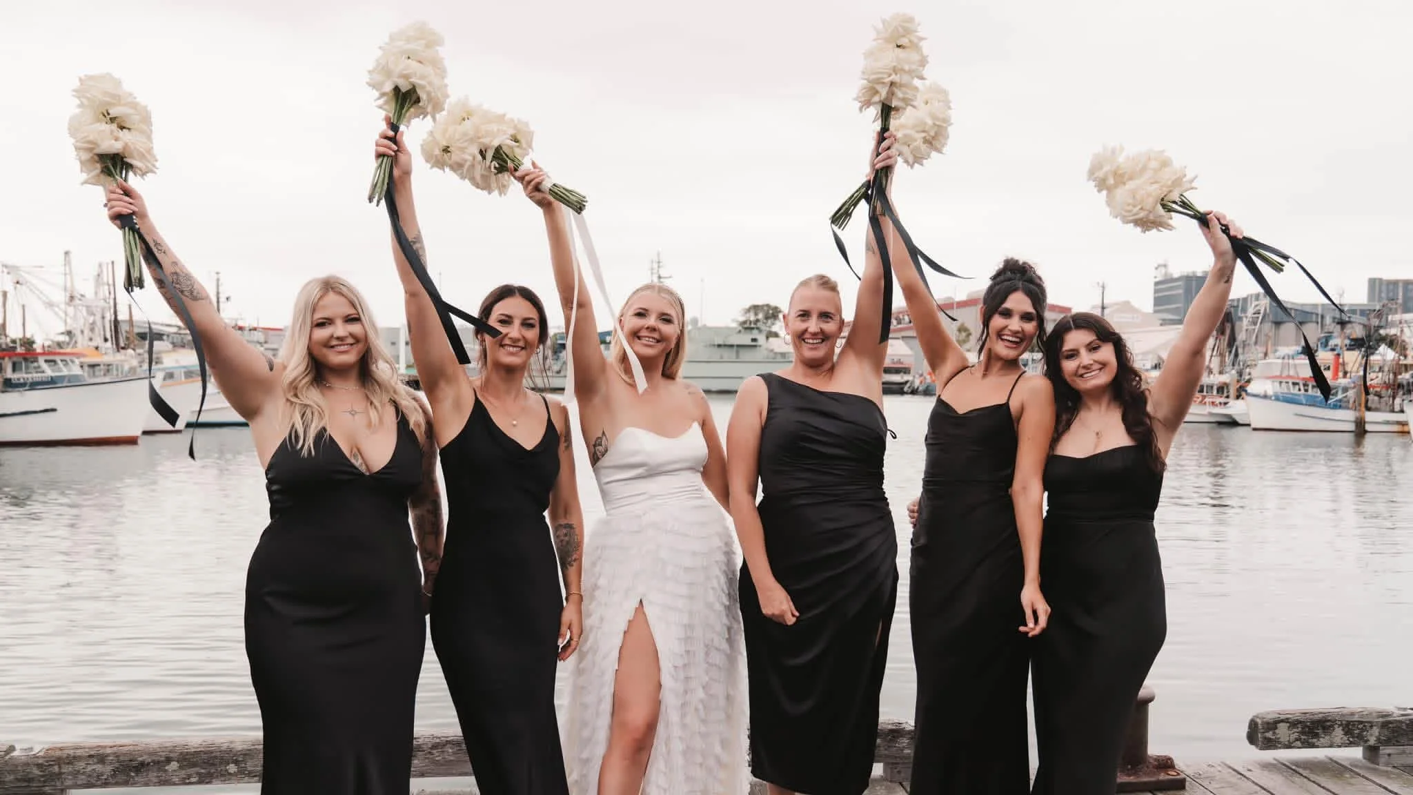 Group of women celebrating with white flower bouquets and wearing black dresses near a marina with boats