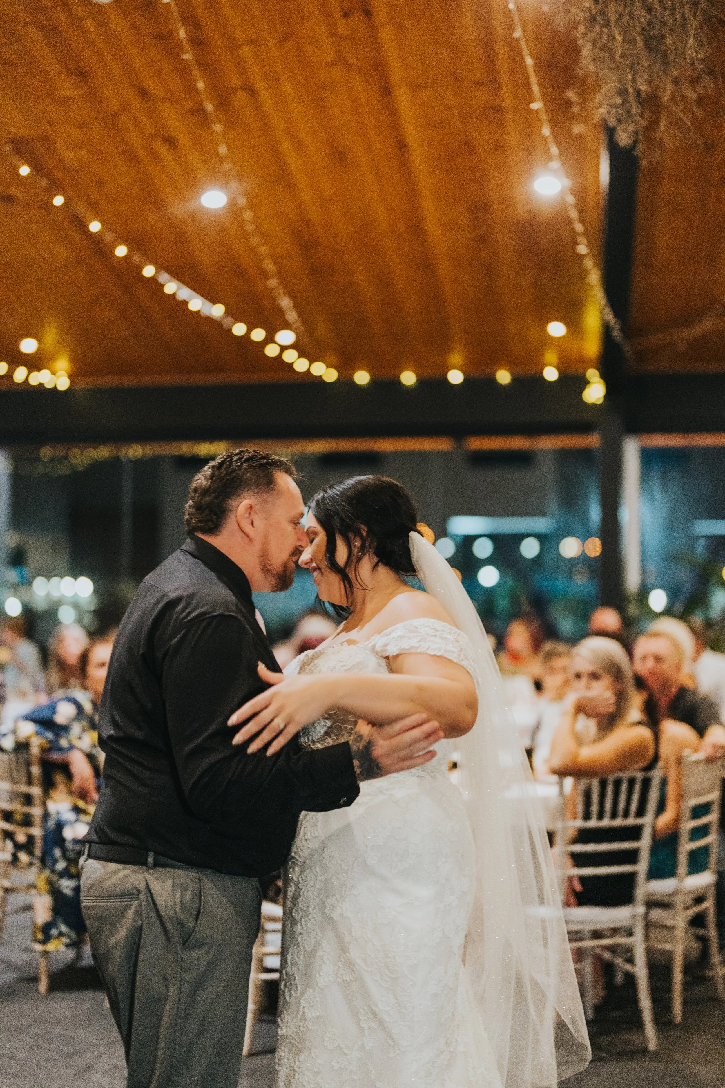A bride and groom share a dance at their wedding reception, with guests seated in the background under a wooden ceiling decorated with string lights.