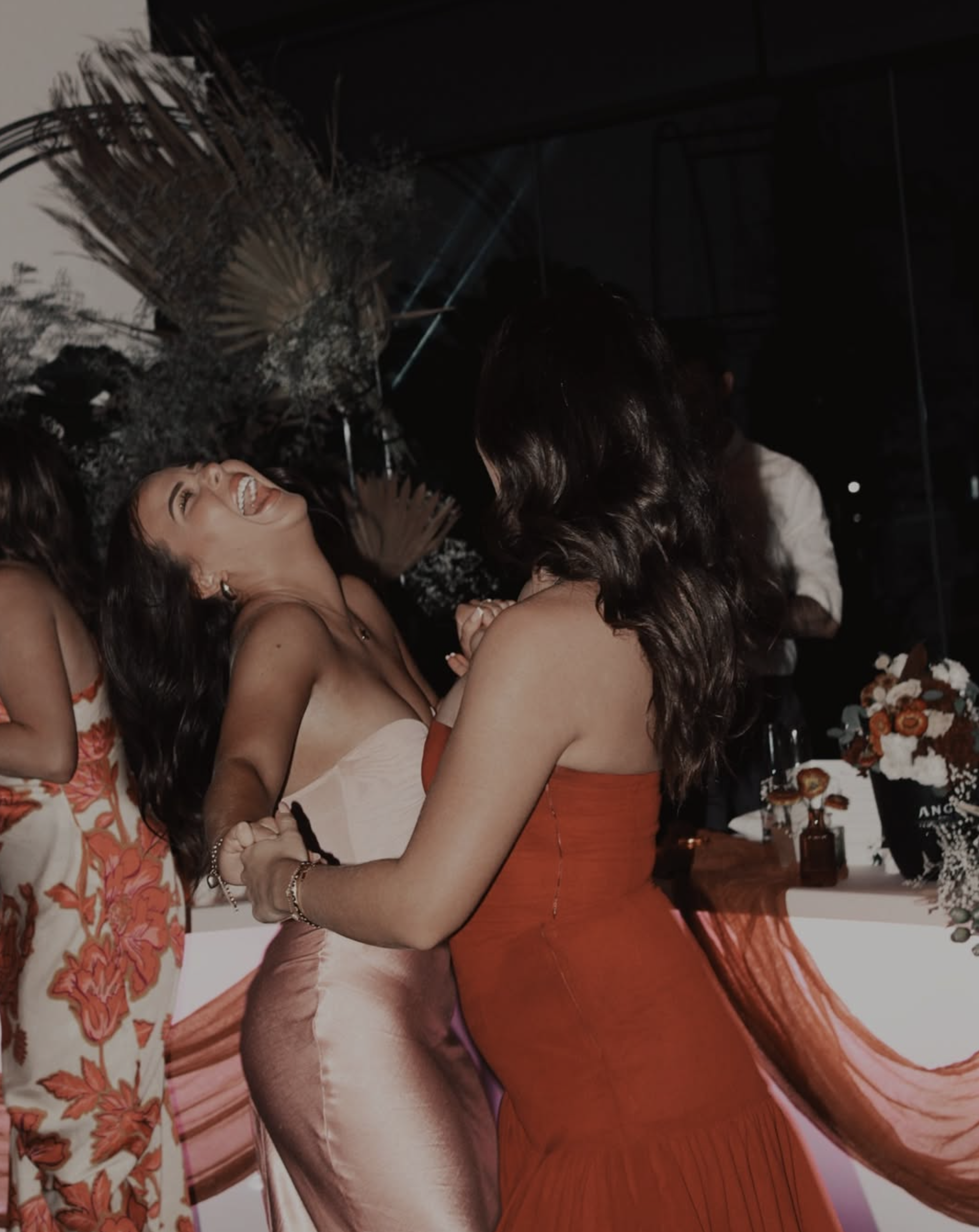 Two women: one in a pink satin dress and the other in an orange dress, joyfully dancing and holding hands at a celebration or party, with a floral centerpiece in the background.
