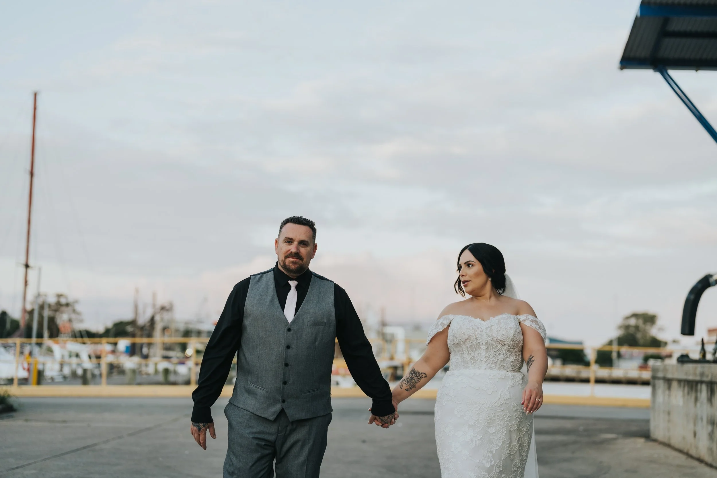 A man and woman in wedding attire walking hand in hand outdoors near a marina with boats, under a cloudy sky.