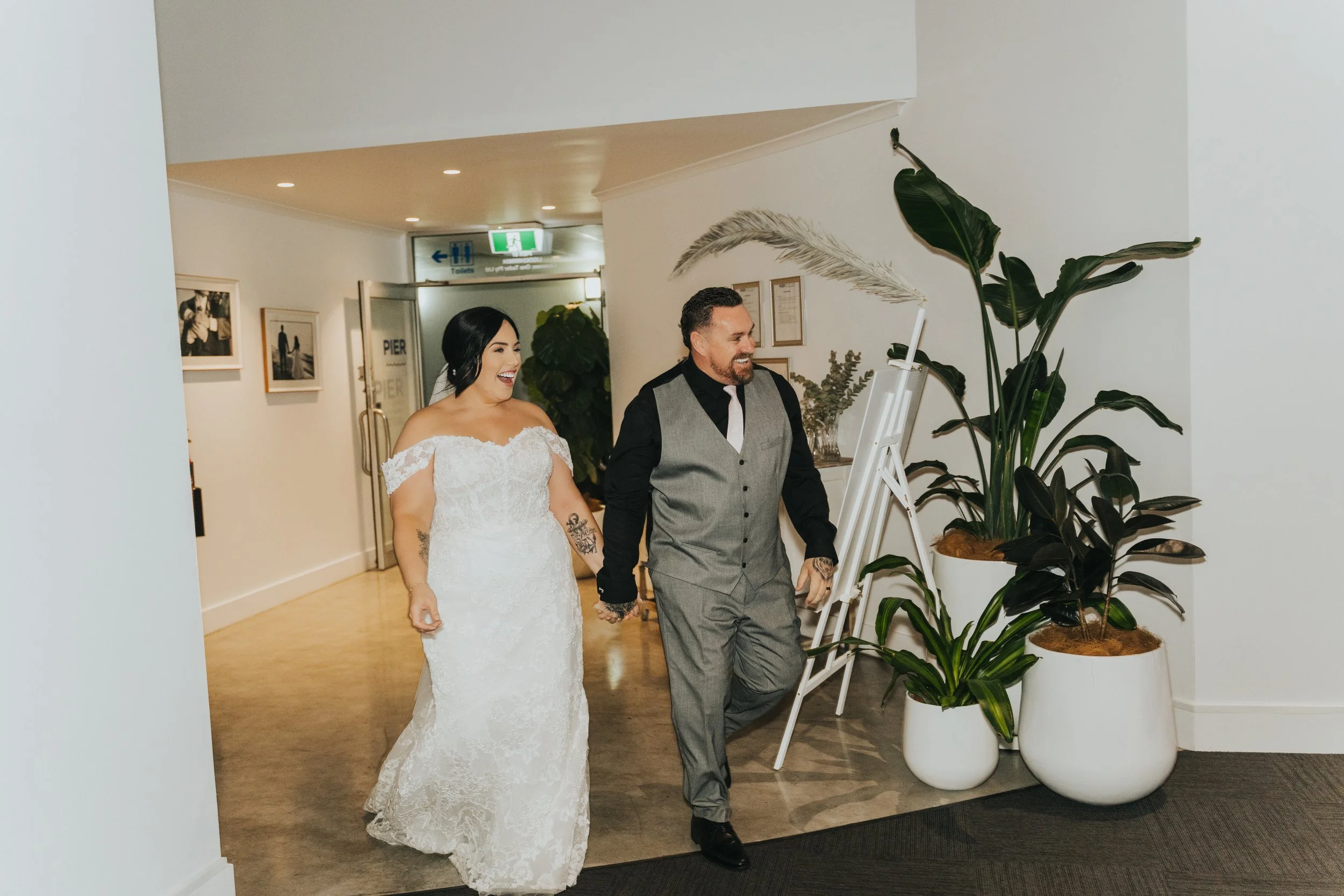 A bride and groom holding hands and smiling while walking through a modern indoor space with potted plants and framed photos on the wall.