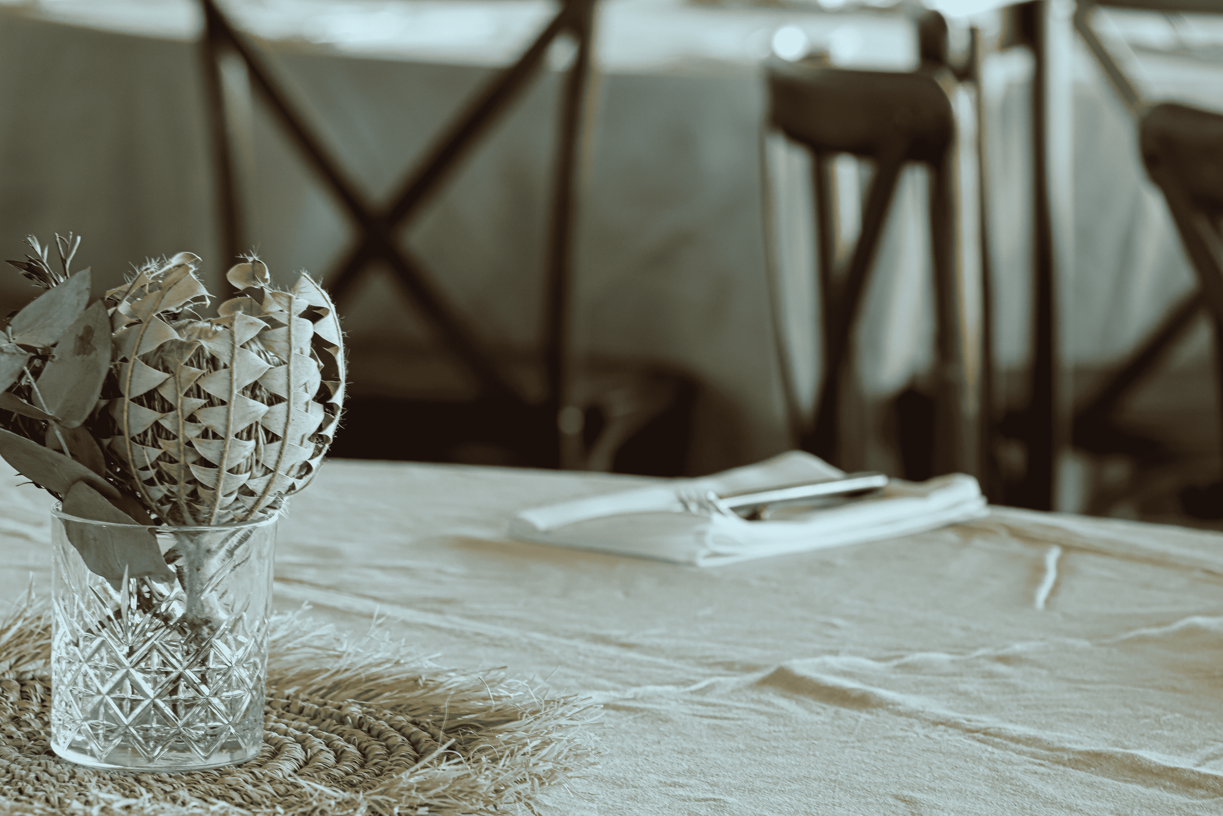 A table with a woven placemat and a glass vase holding preserved flowers, with chairs and a window in the background.