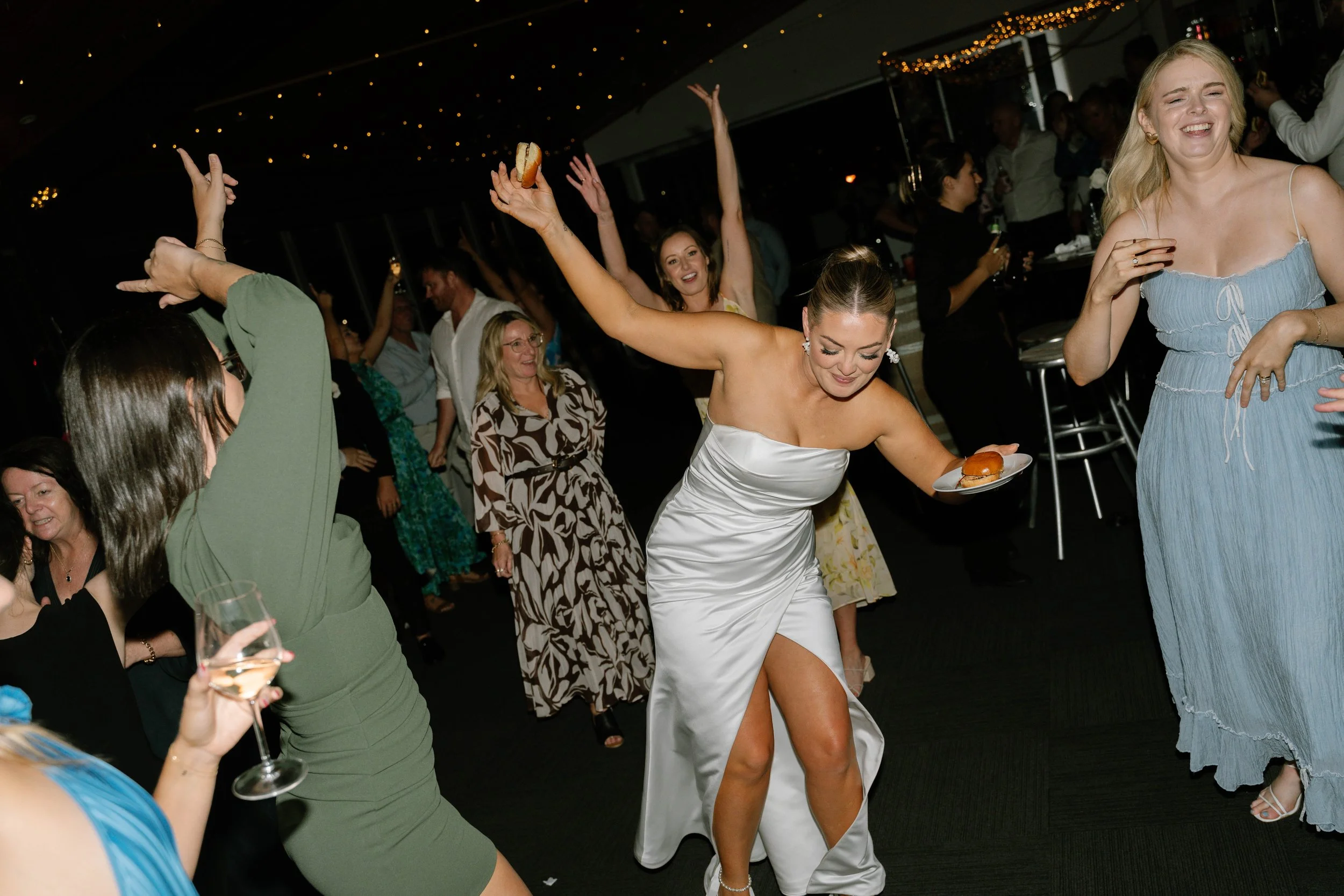 A woman in a white dress dancing at a celebration, holding a plate with a hot dog, surrounded by other women and guests enjoying the event, under warm string lights.