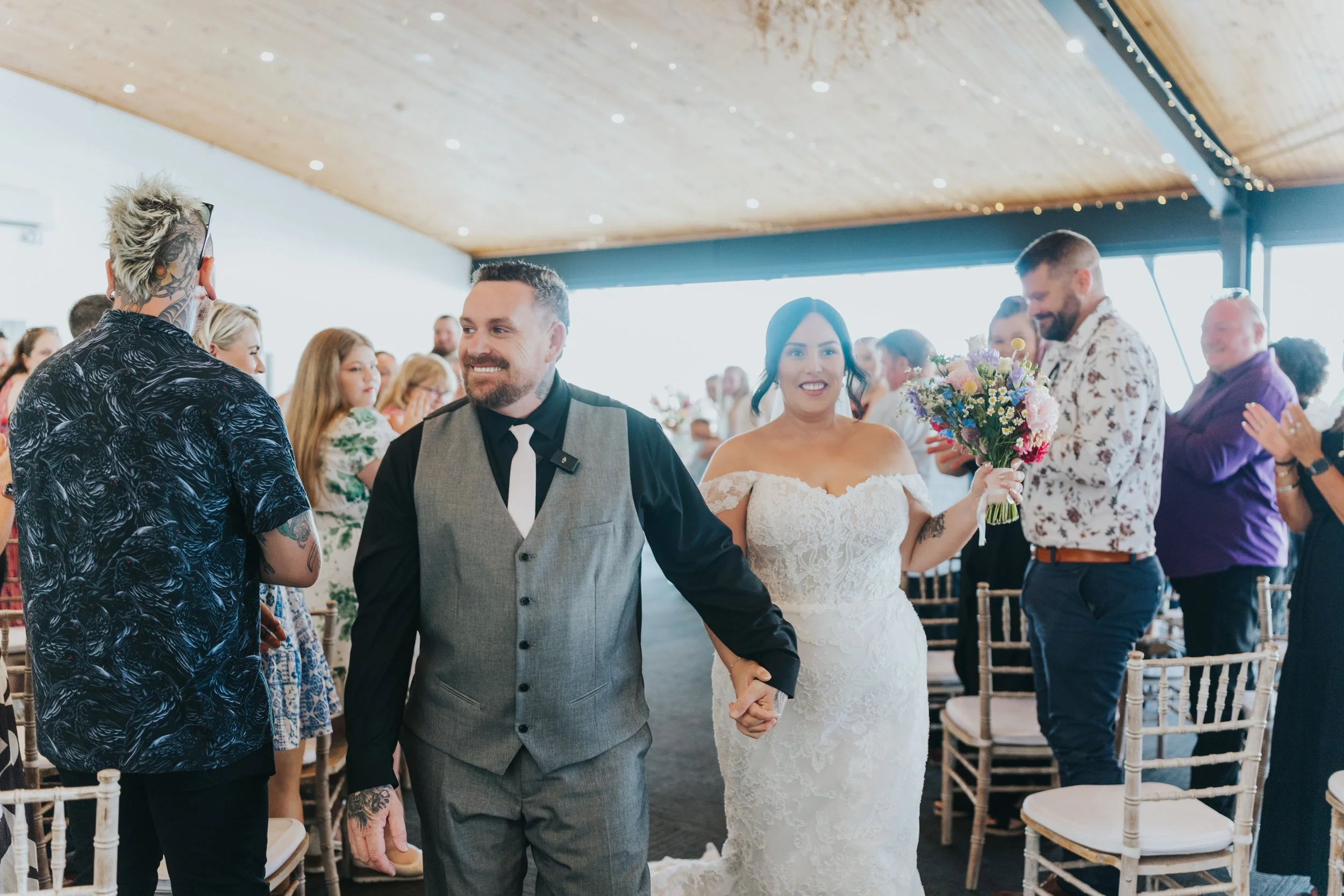 A newlywed couple walking hand in hand down the aisle at their wedding ceremony, surrounded by guests clapping and smiling, inside a decorated venue with a wooden ceiling and string lights.