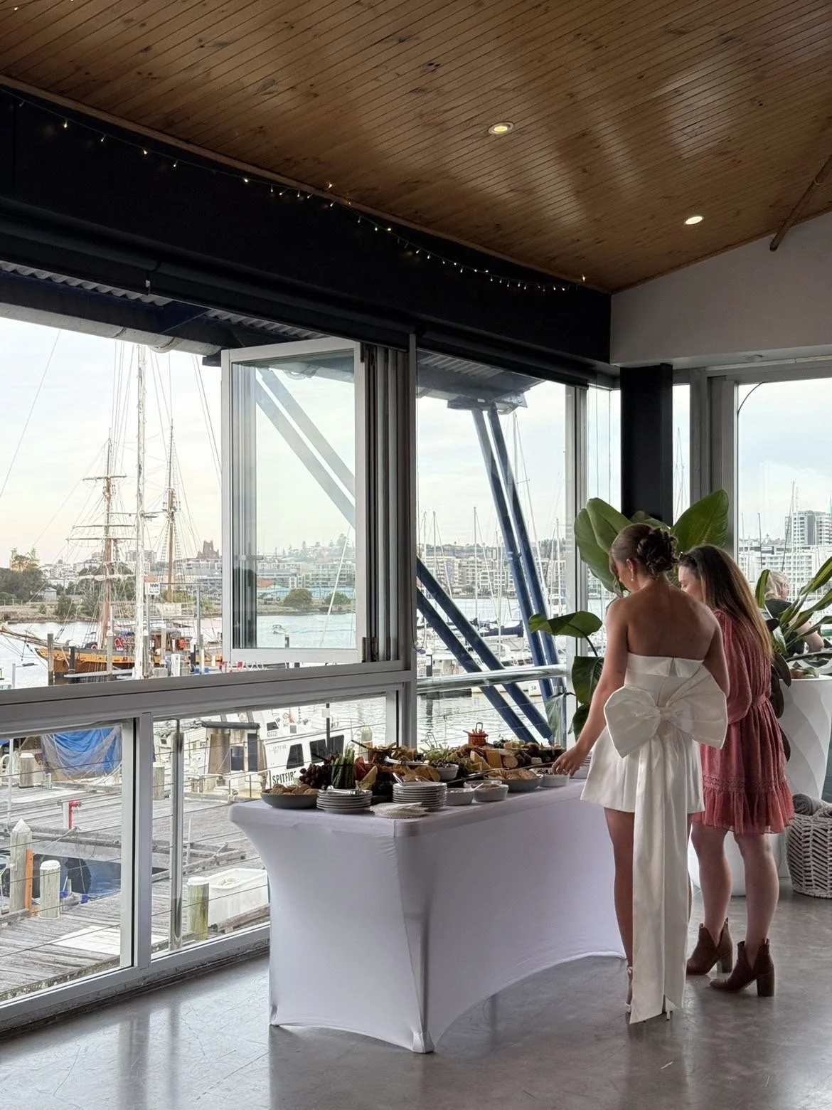 Two women at a buffet table overlooking a marina with sailboats, interior with wooden ceiling and large windows.