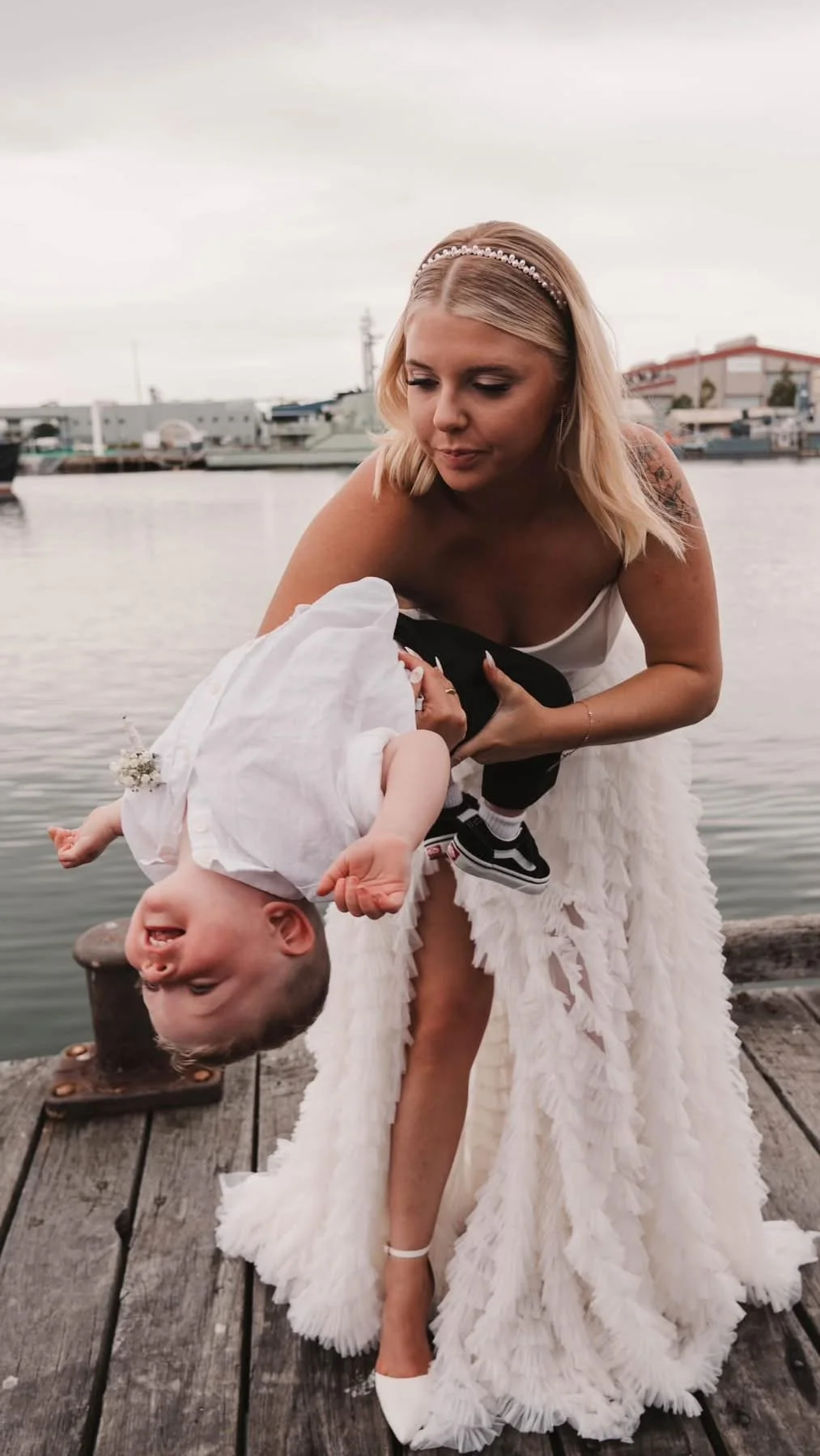 A woman in a wedding dress is holding a laughing young boy upside down on a wooden dock by the water, with boats and buildings in the background.