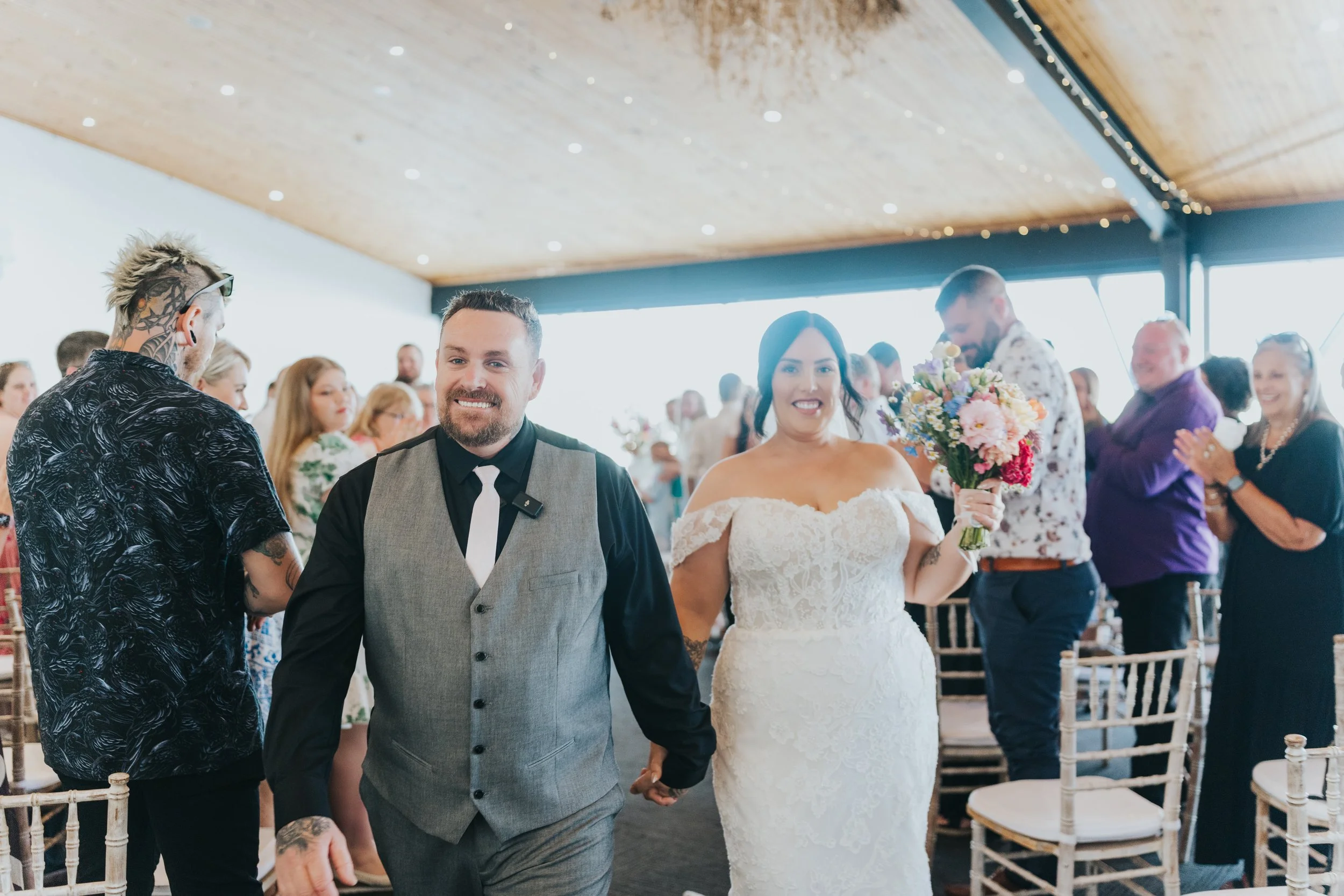 A wedding ceremony with a bride in a white lace dress holding a bouquet of flowers, and a groom in a gray vest and black shirt, walking hand in hand down the aisle, surrounded by seated guests.