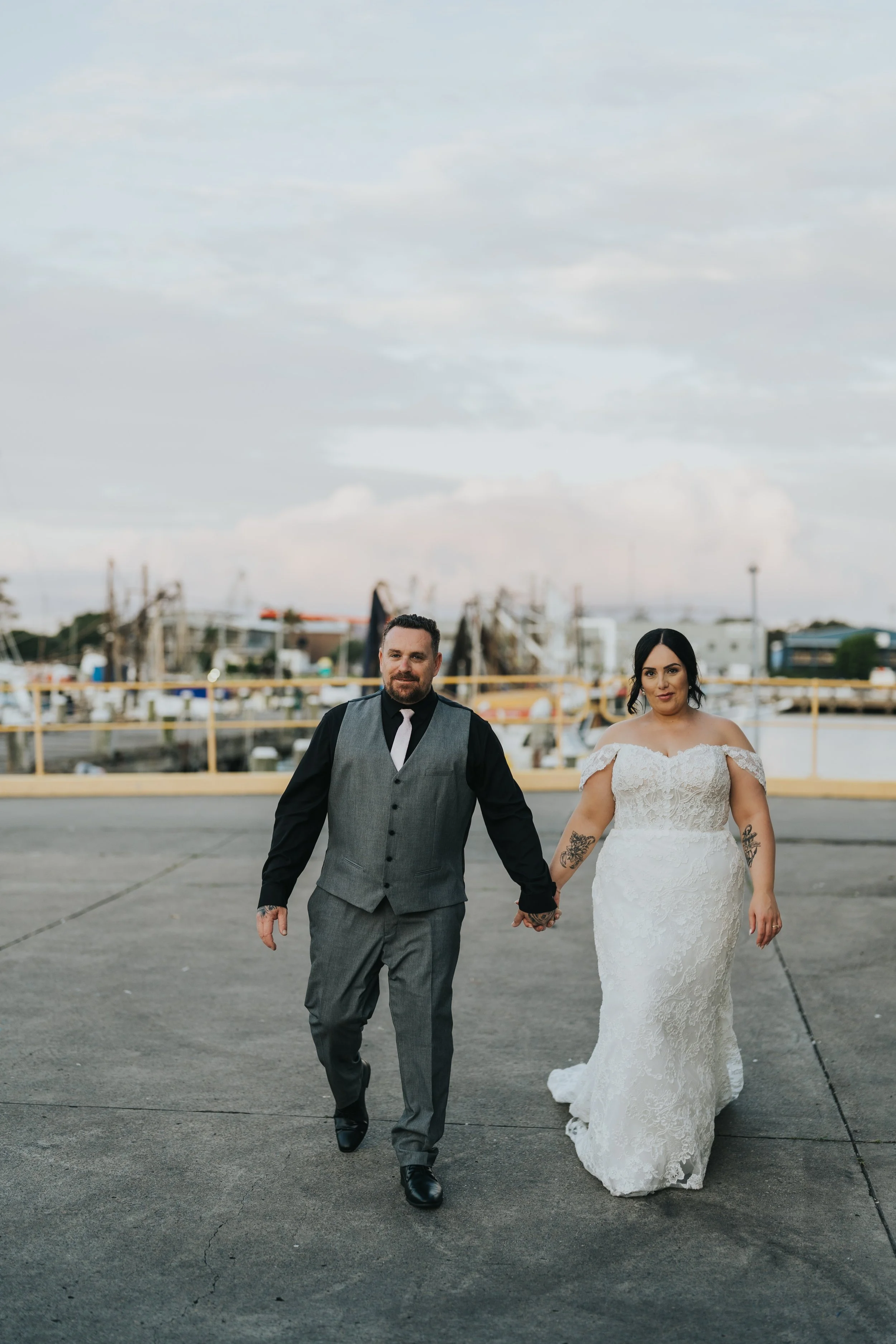 A newlywed couple walking hand in hand outdoors near a marina with boats in the background. The groom wears a gray vest and black shirt, and the bride wears a white lace off-shoulder wedding dress.