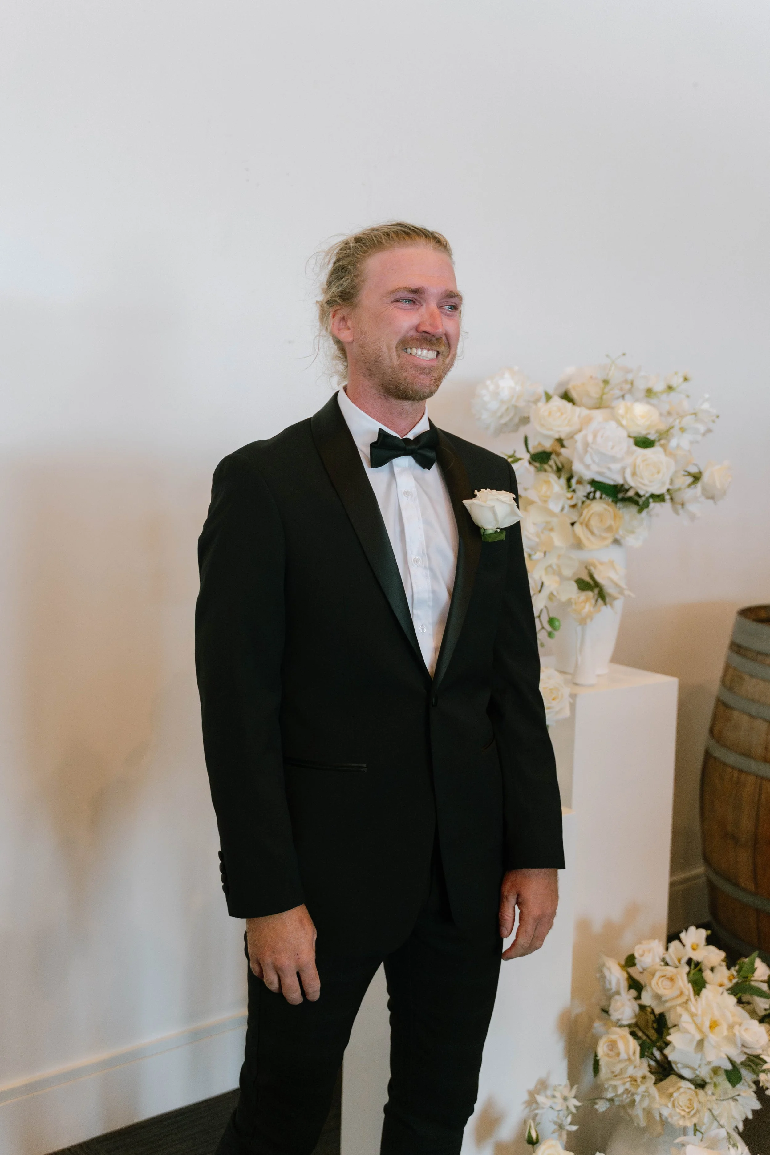 A man in a black tuxedo and bow tie smiling at a formal event, with flowers in the background.