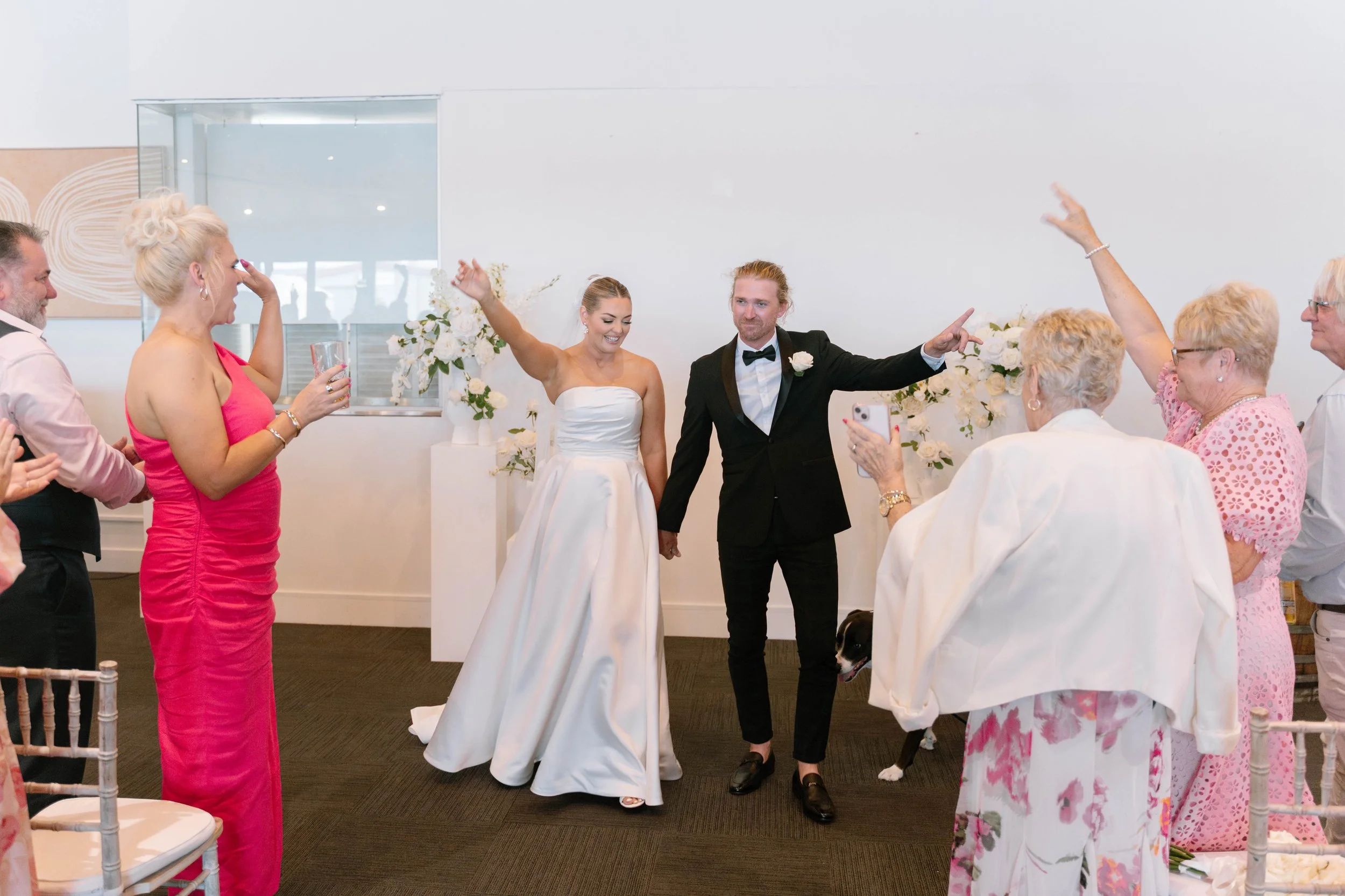 Bride and groom holding hands and celebrating with family and friends at a wedding ceremony, with floral decorations and a black dog present.