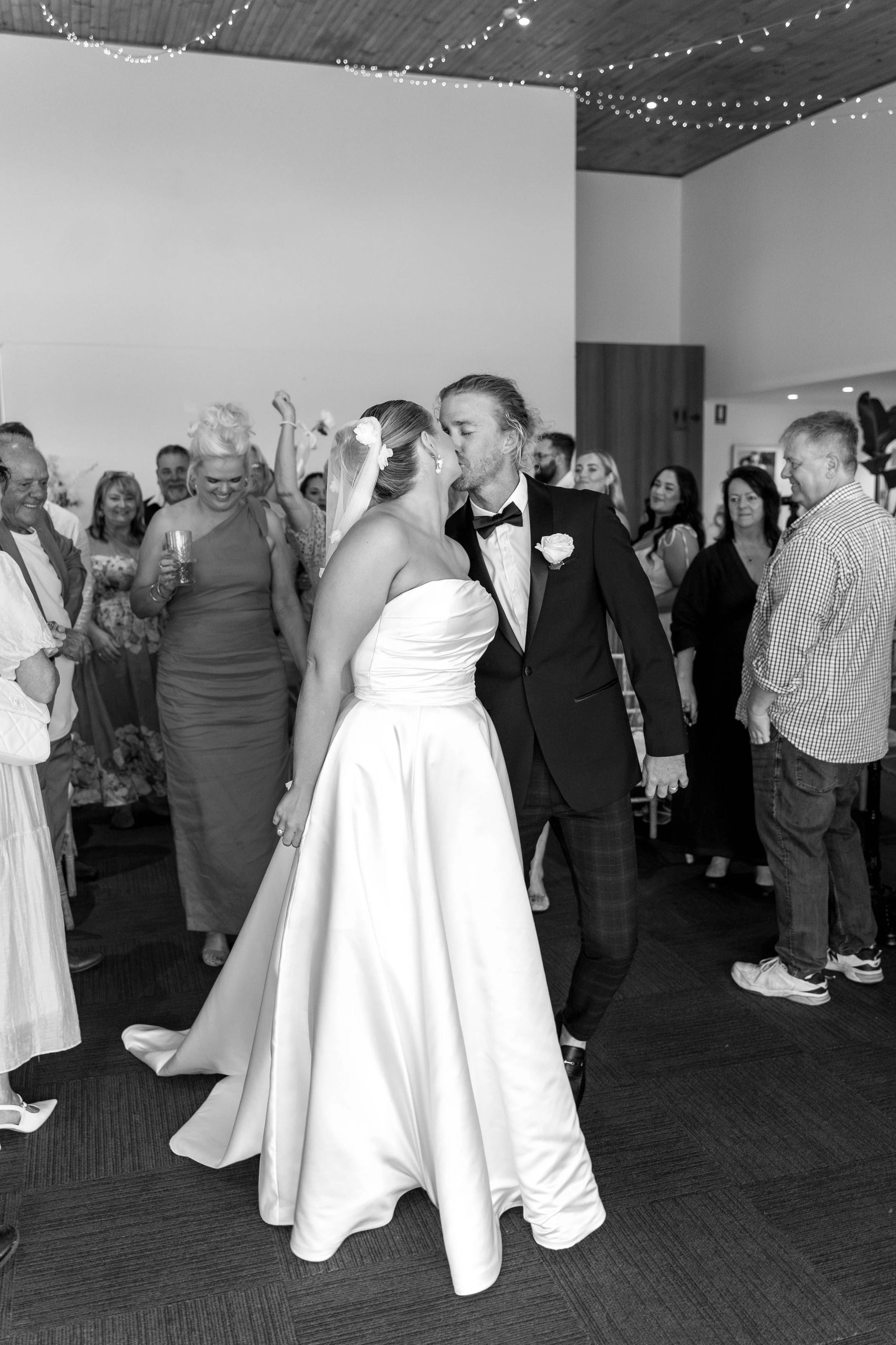 Black and white photo of a bride and groom kissing, surrounded by wedding guests at their reception.