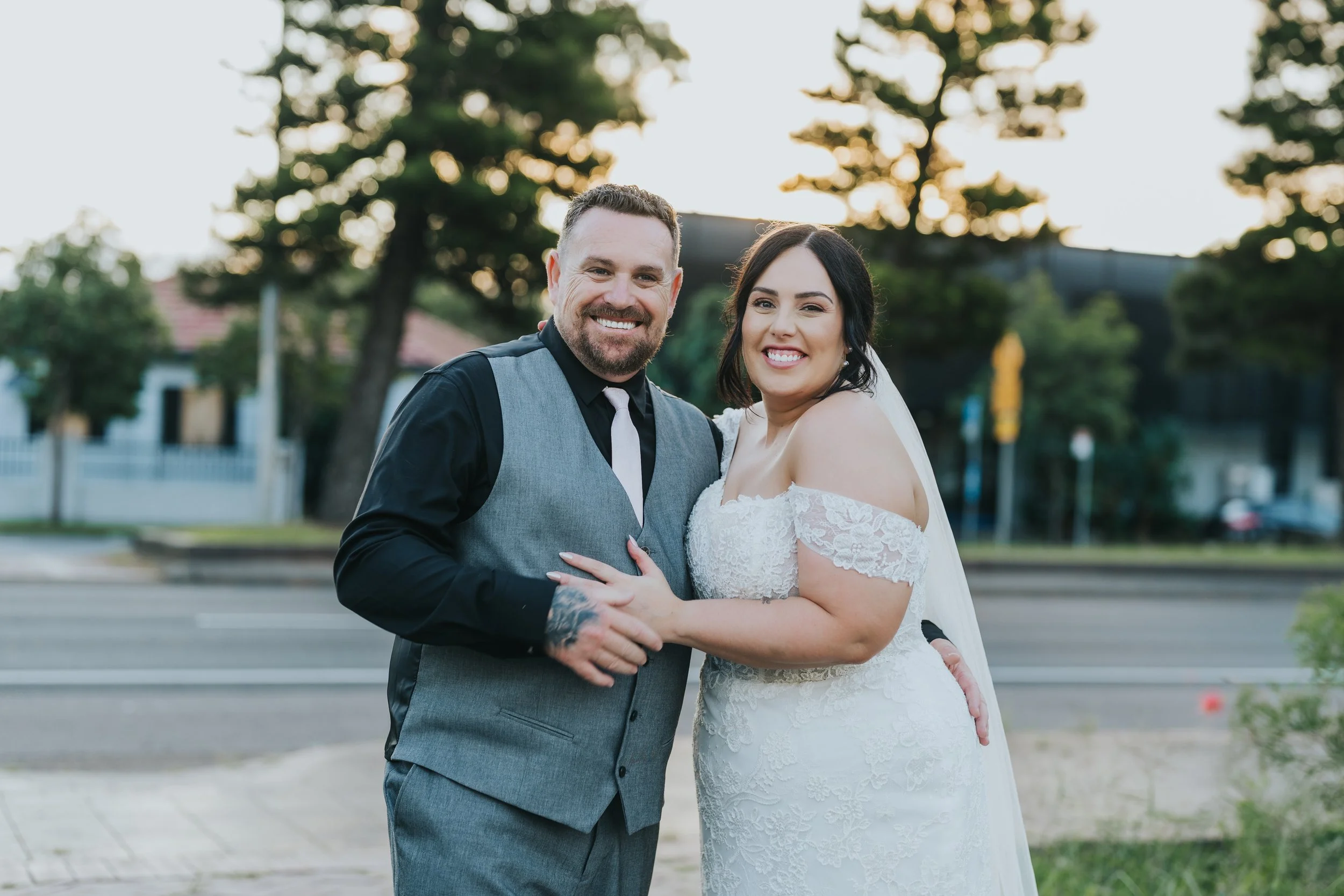 A newlywed couple smiling and holding hands outdoors during sunset, with trees and houses in the background.