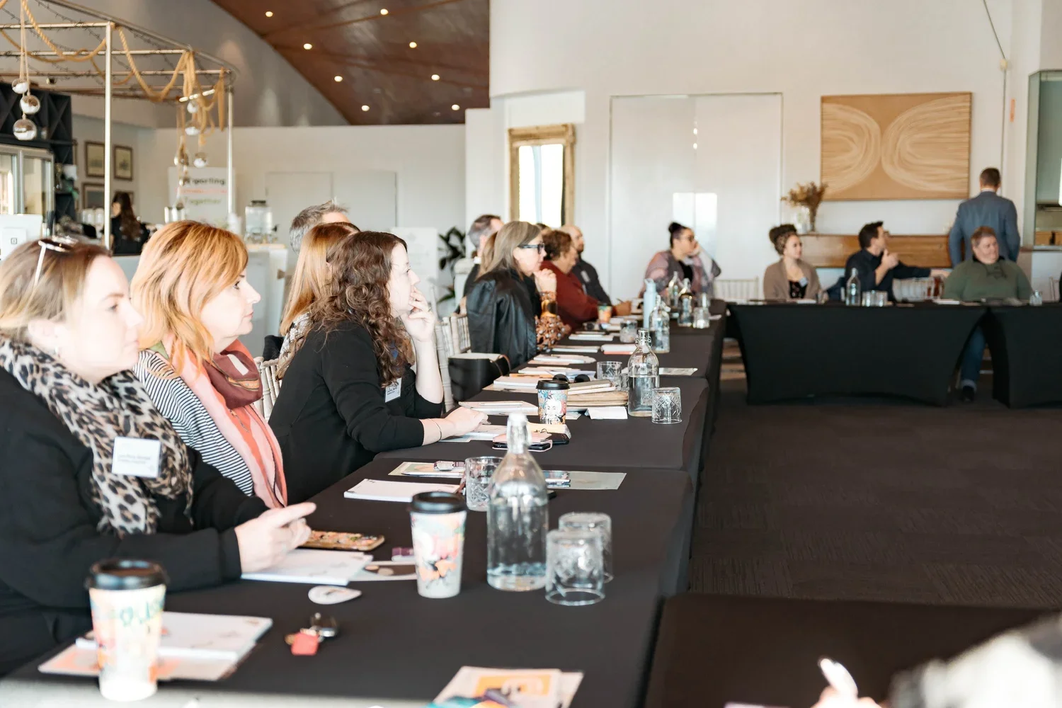 People sitting at a long table in a conference room, listening to a speaker at the front.