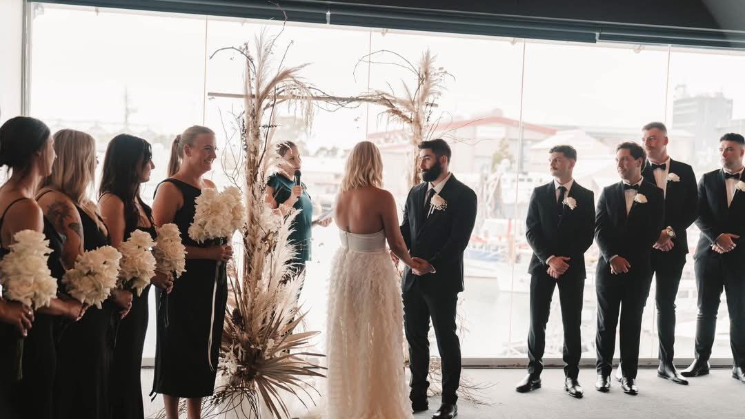 A wedding ceremony with a bride and groom exchanging vows, surrounded by bridesmaids and groomsmen in formal black suits, near large windows overlooking a marina with boats.