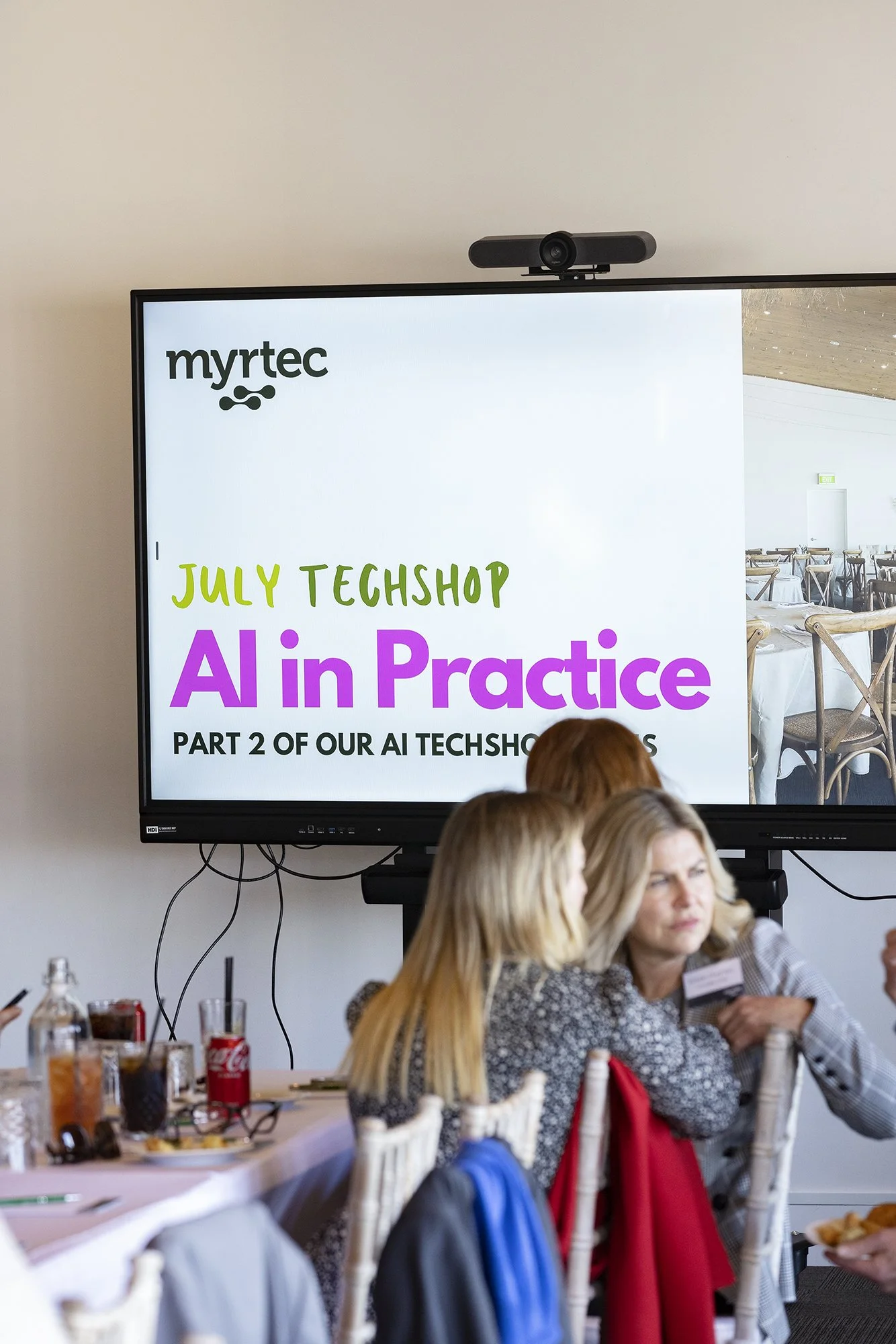 Large screen displaying a presentation for an AI tech event titled 'July Techshop: AI in Practice' with people seated in front of the screen.