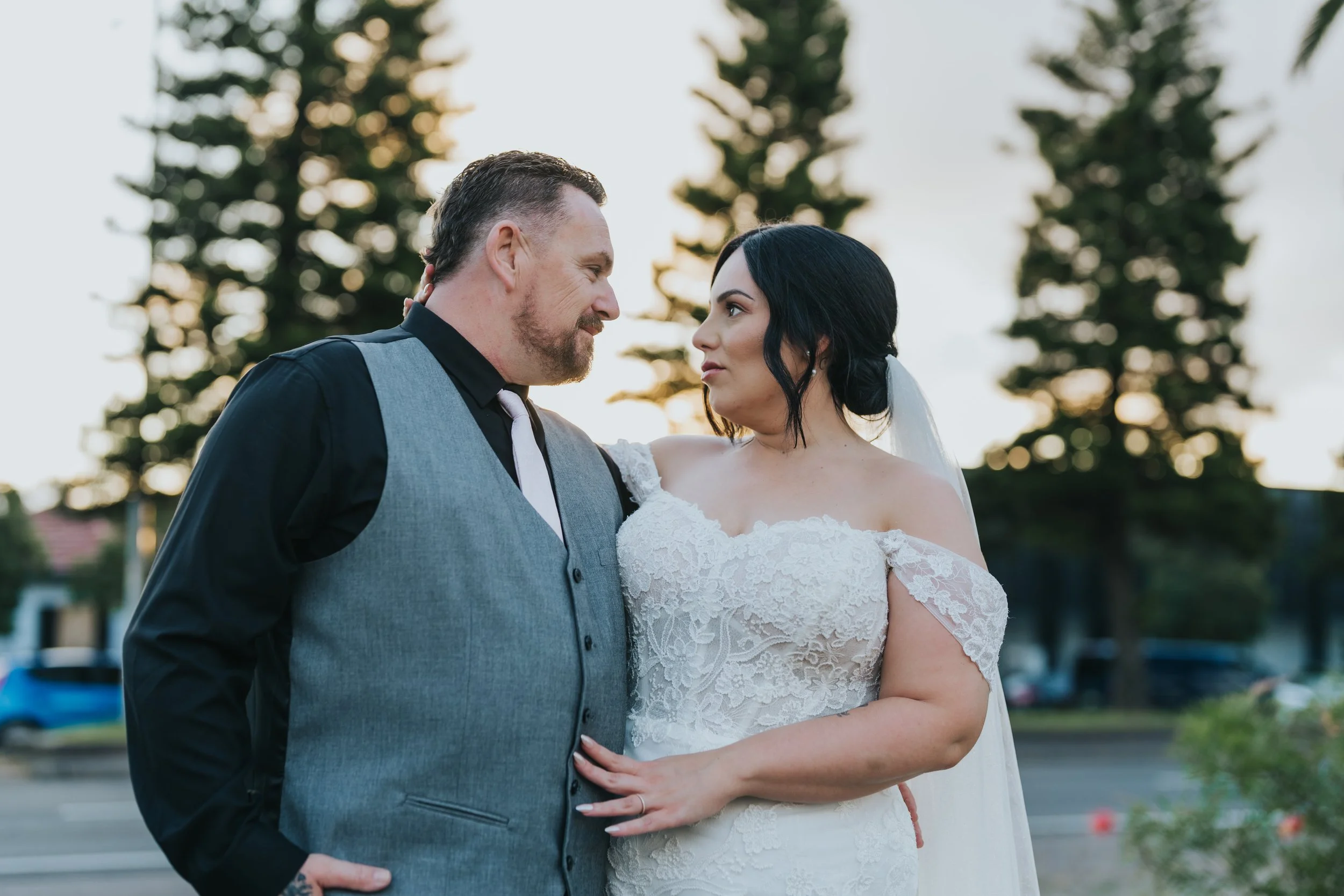 A bride and groom touching foreheads outdoors during sunset, with trees and parked cars in the background.