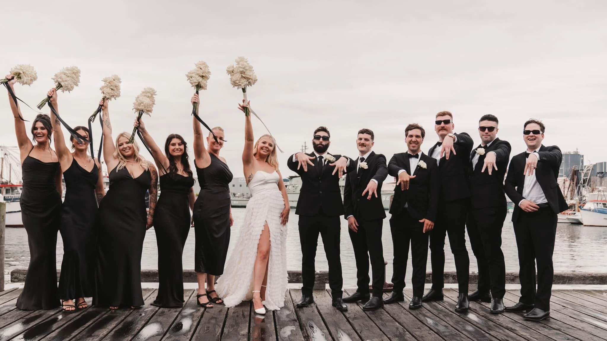 A wedding party on a dock by the water, with eight women in black dresses holding white bouquets and four men in black suits and ties, posing with playful hand gestures.