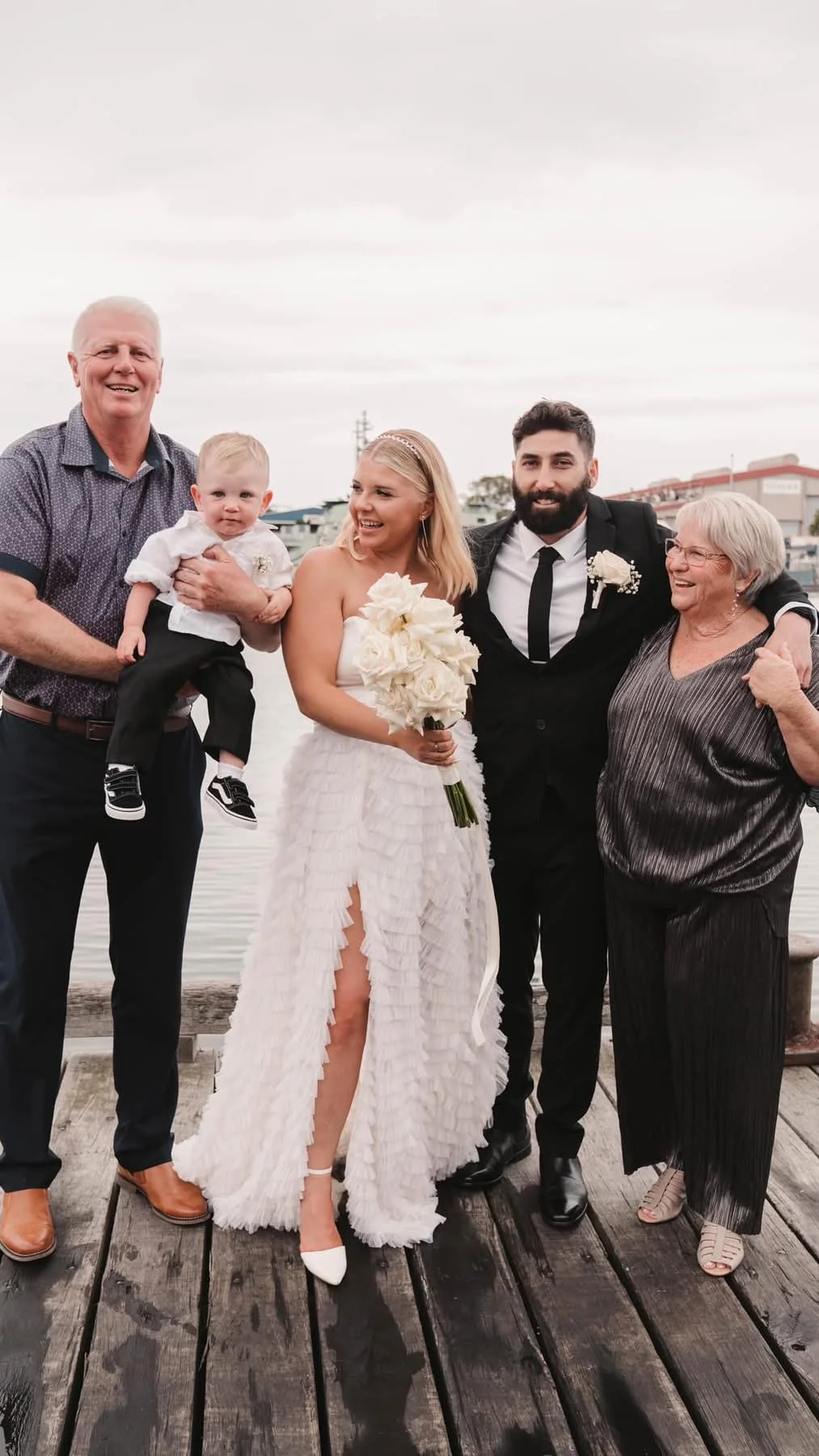 People celebrating a wedding on a wooden dock by the water, including a bride in a white wedding dress holding a bouquet, groom in a black suit, two older women, and a man holding a young child, overcast sky in background.