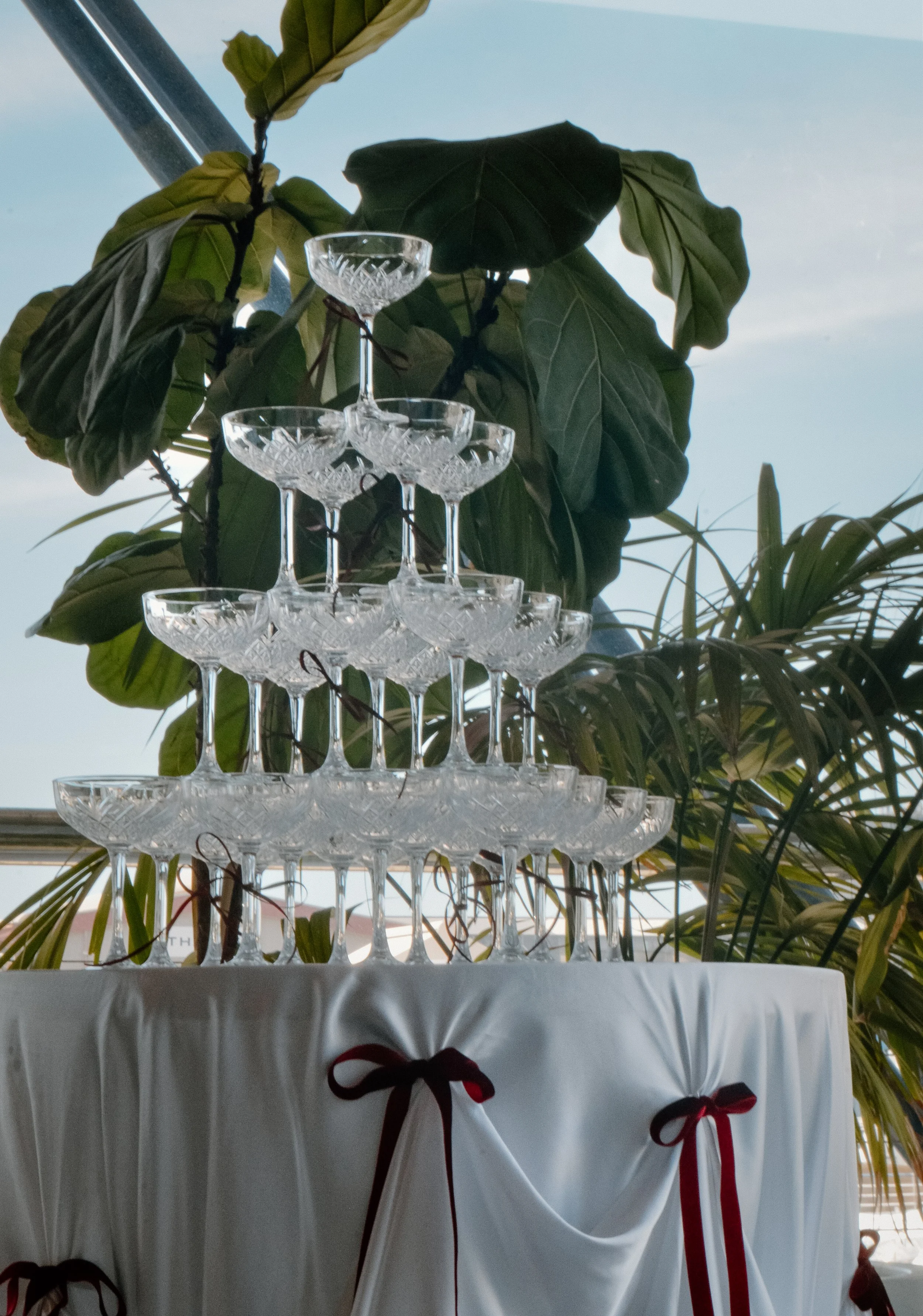 A pyramid of stacked crystal champagne glasses on a table draped with white cloth, decorated with red ribbons, in front of green leafy plants and a blue sky.