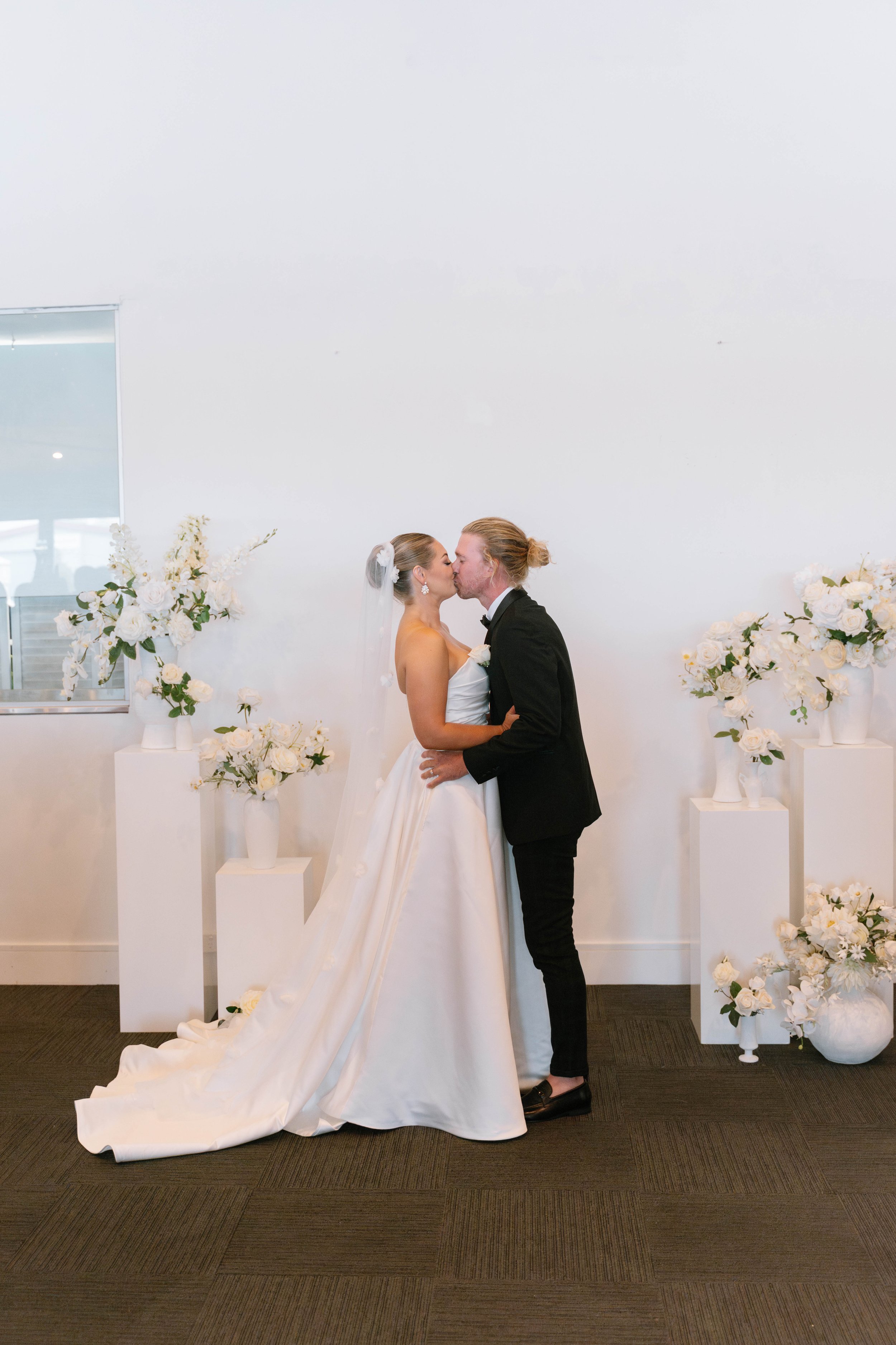 A bride and groom kissing at their wedding ceremony indoors, surrounded by white floral arrangements in vases on pedestals, with a white wall in the background.