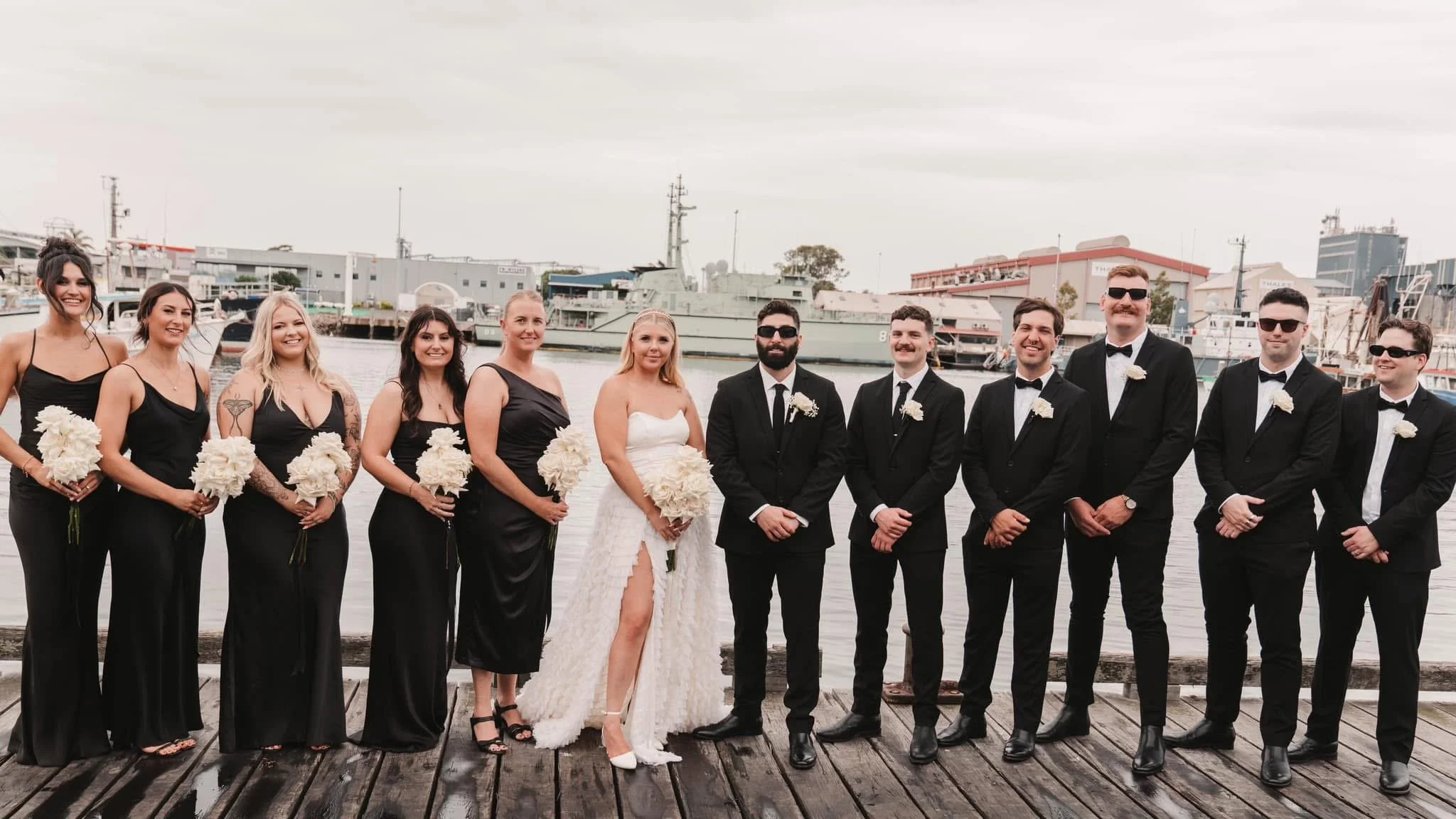 A wedding party standing on a wooden dock near water with ships in the background. The group consists of women in black dresses and men in black suits and sunglasses, with the bride in a white dress and the groom in a black suit. They are smiling and