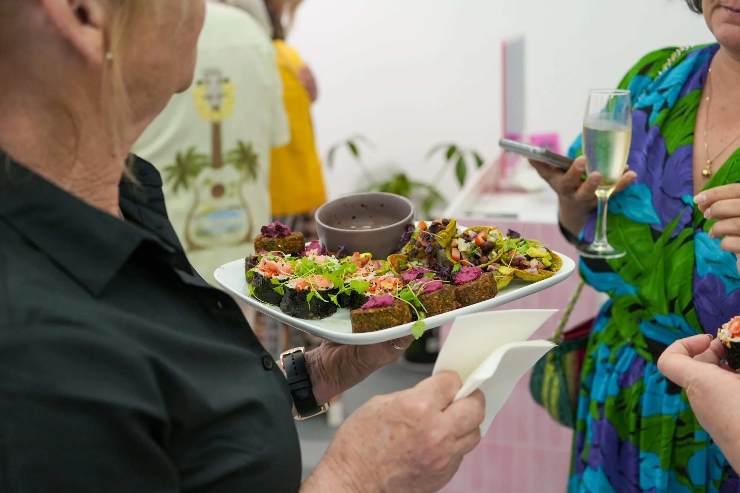 A woman holding a plate of assorted colorful appetizers, including small bites with greens, purple toppings, and crispy items, at a social gathering with others holding drinks.