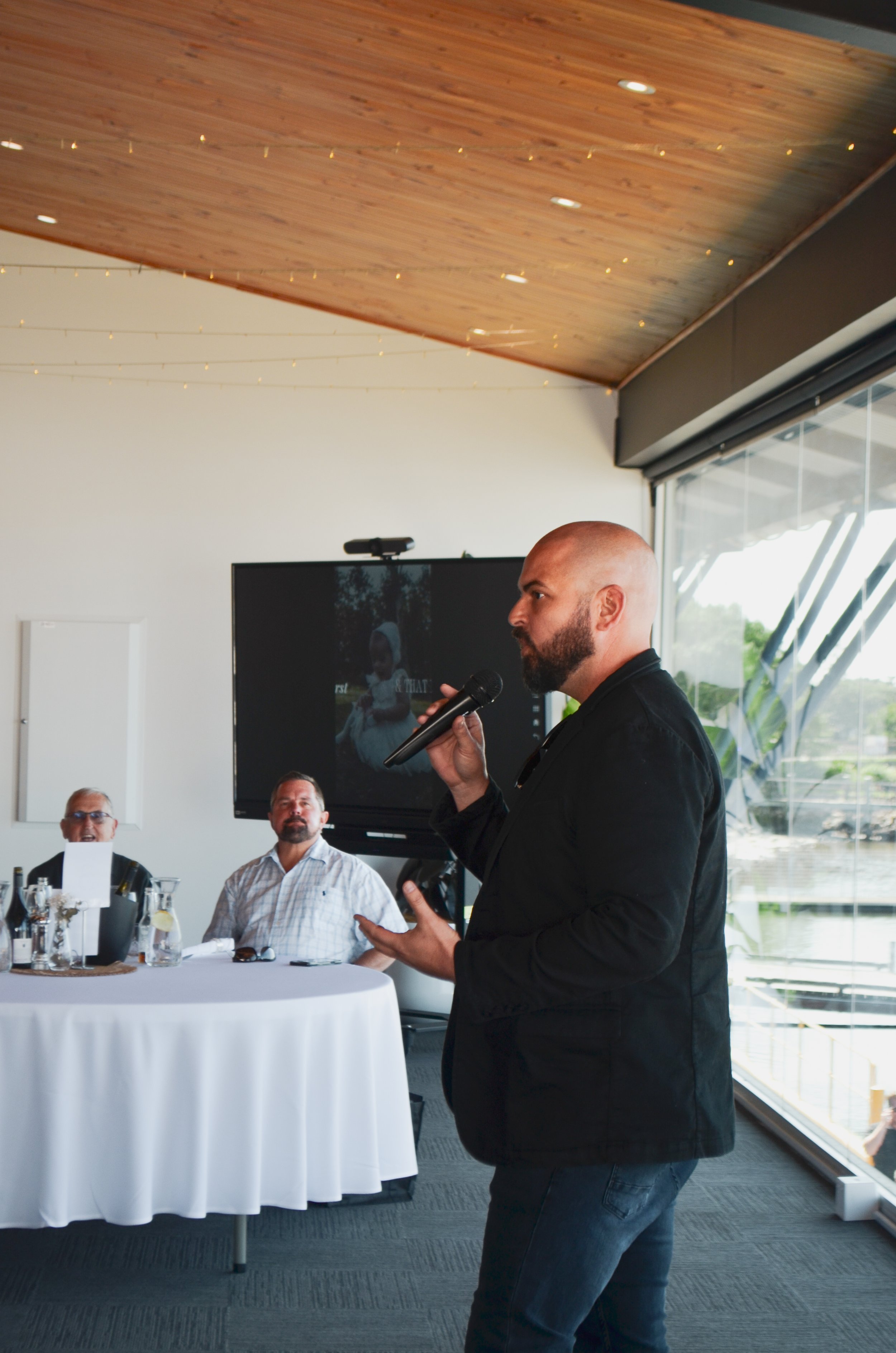 A man with a beard and shaved head holding a microphone and speaking at a formal event. Two men are seated at a round table with white tablecloths in the background, in a room with a wooden ceiling, large windows, and a television screen.