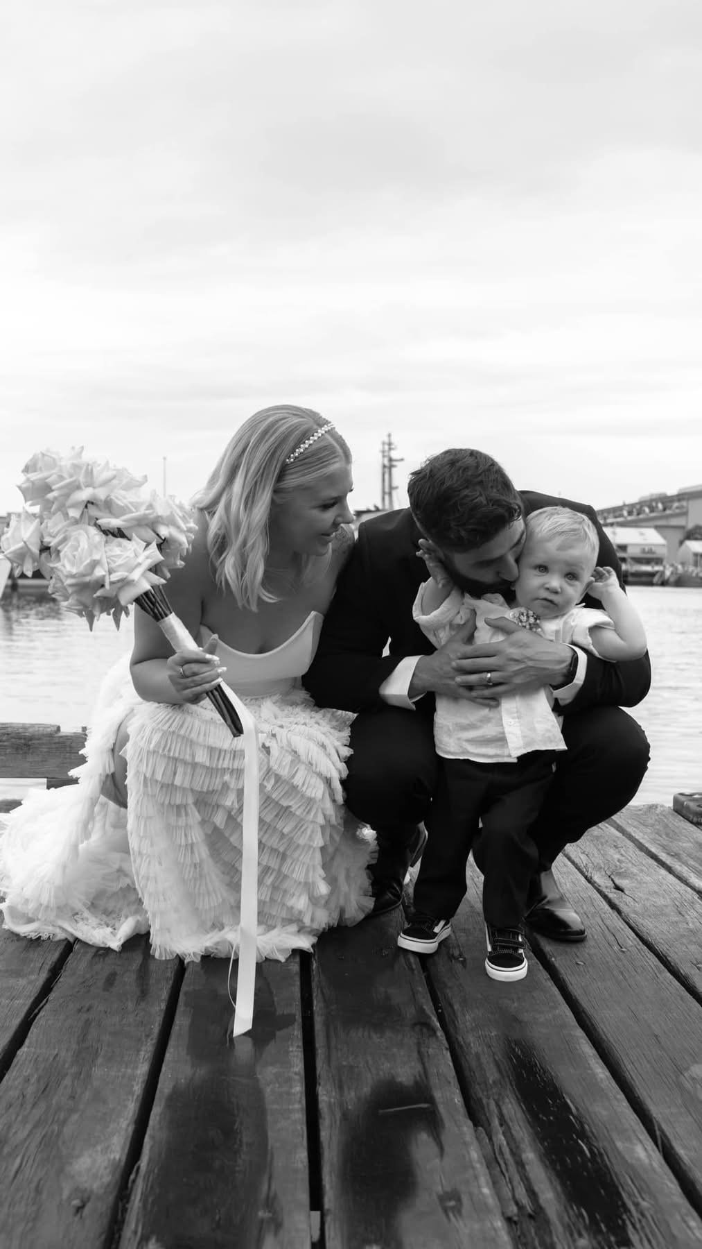 Black and white photo of a newlywed couple with a young child on a wooden pier near water, with boats and buildings in the background. The bride holds a bouquet of flowers, and the groom kisses the child.