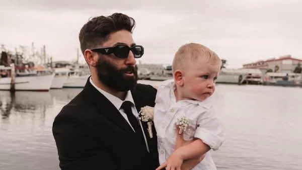 A man dressed in a black suit and sunglasses holding a young child with blonde hair by the waterfront.
