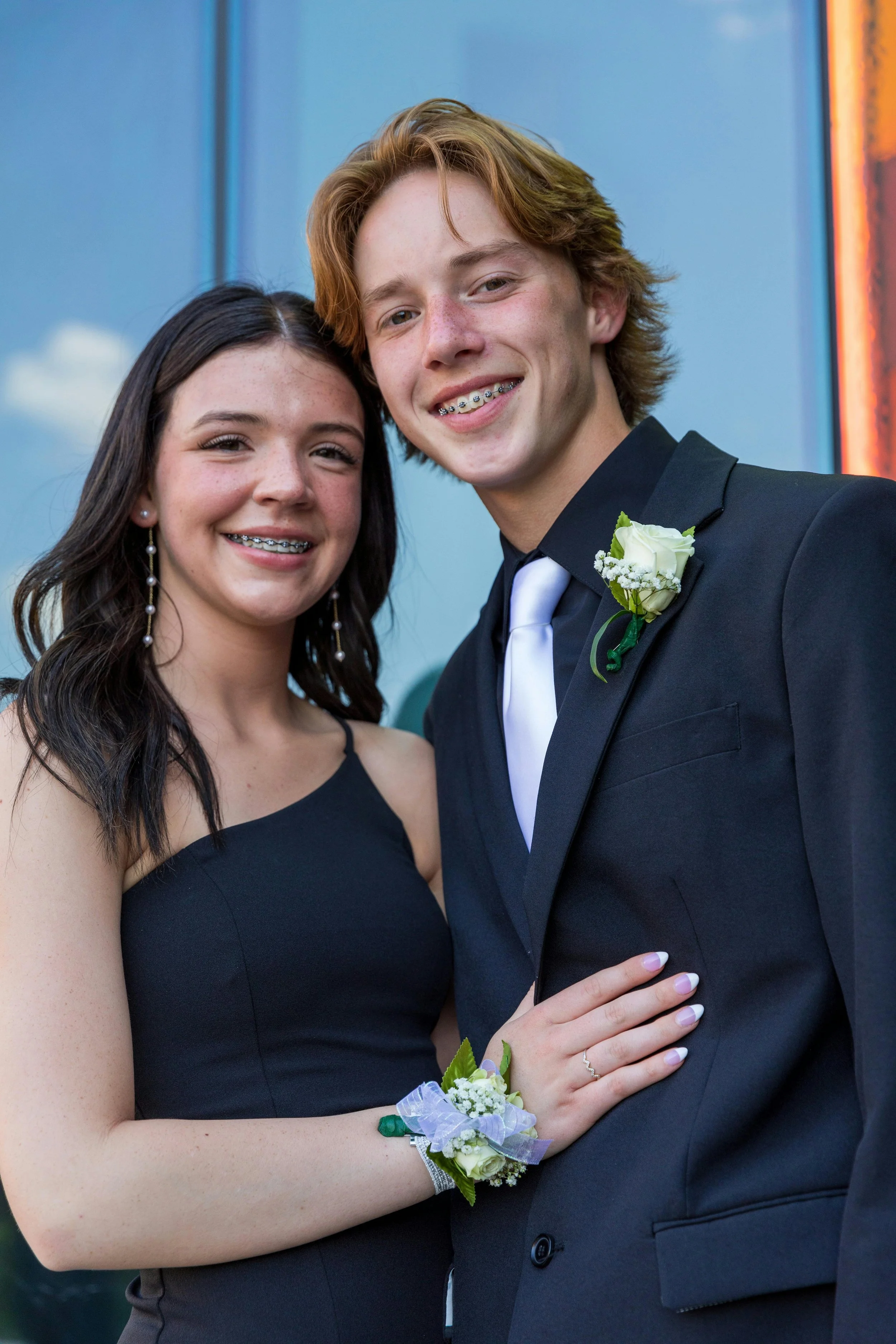 A young couple dressed in formal attire, with corsages and boutonnieres, smiling for a photo at a prom or special event.