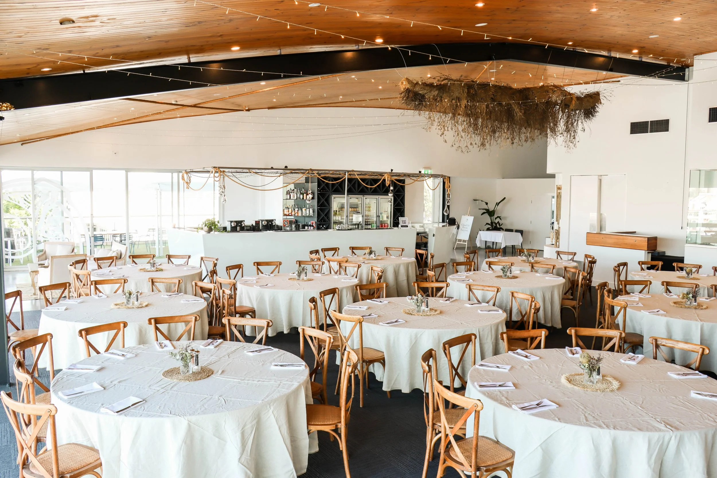 A decorated banquet hall with round tables covered in white tablecloths, each set with folded napkins and small flower arrangements. The hall has a wood-paneled ceiling with hanging string lights and a bar area at the back with bottle shelves and a refrigerator. Natural light fills the room through large windows and sliding doors.