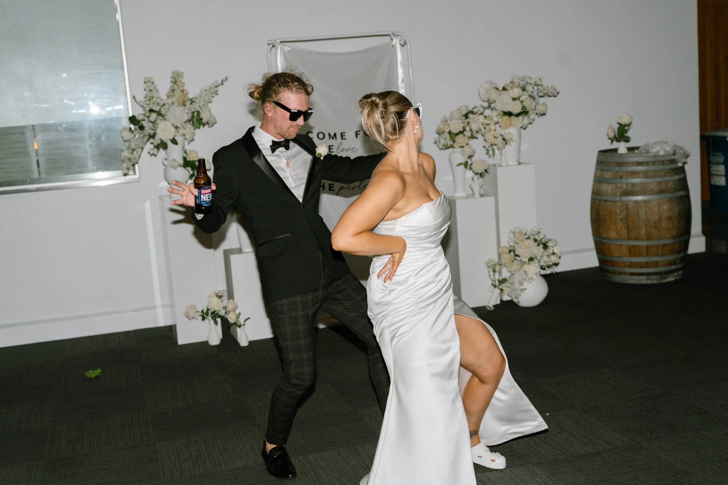A man in a black tuxedo and a woman in a white satin strapless dress dancing at a wedding reception, with floral decorations and a wooden barrel in the background.