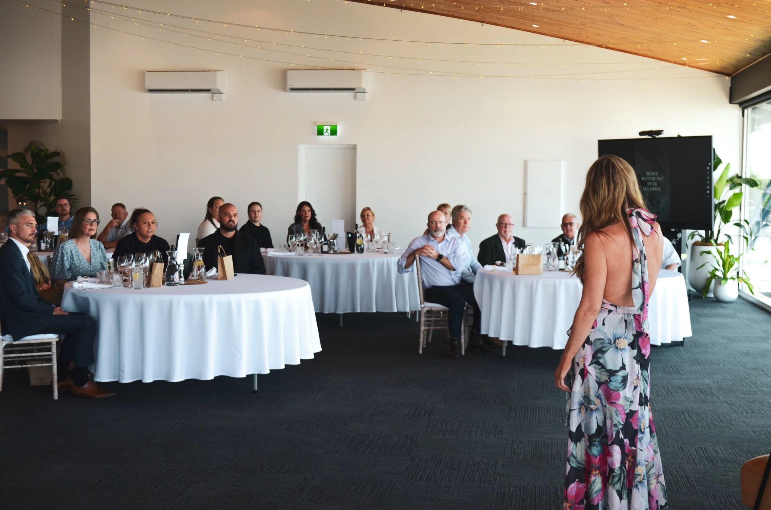 A woman in a floral dress standing in front of a seated audience in a bright, modern room during a presentation.