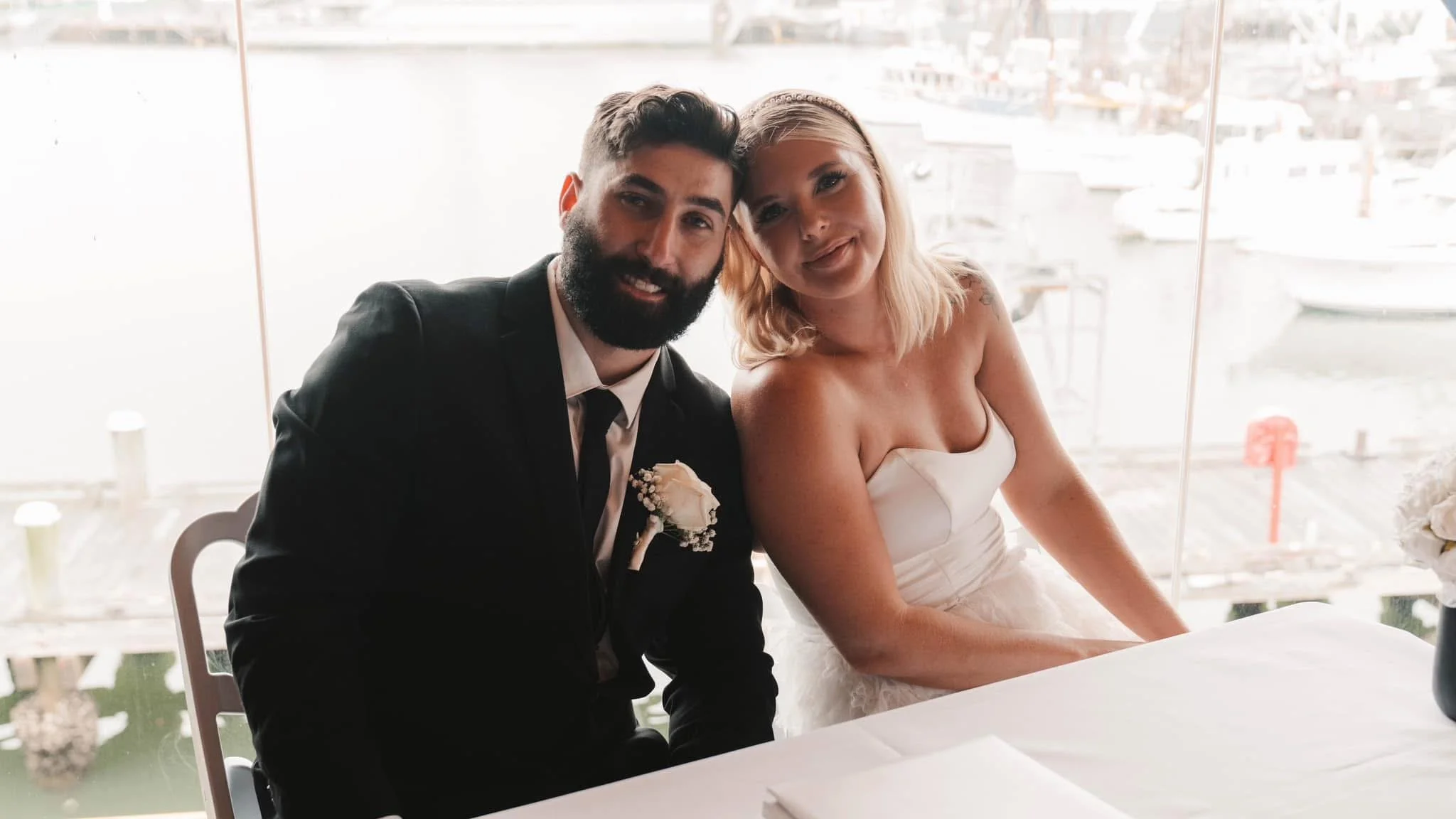 A man in a dark suit and a woman in a white dress sitting together at a table during a wedding, with a marina and boats visible through large windows behind them.