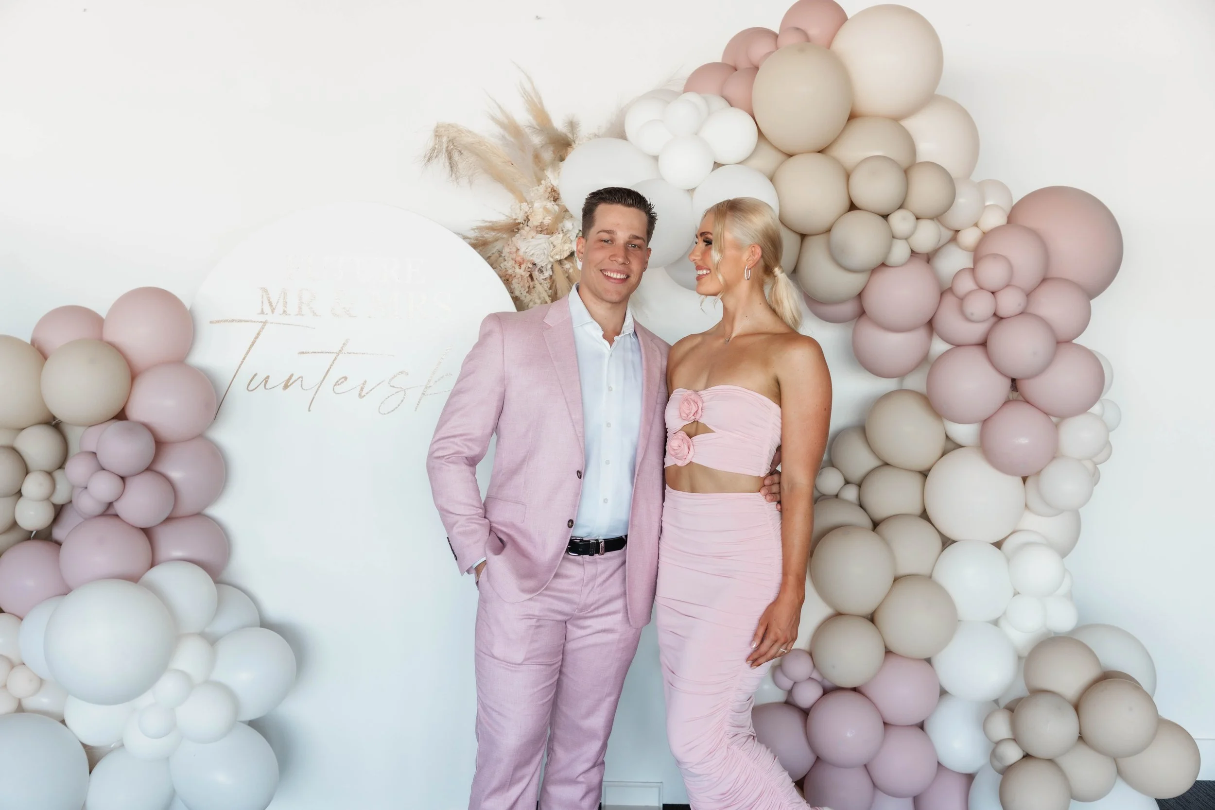 A man and woman dressed in pastel pink outfits stand together in front of a balloon backdrop. The man wears a pink suit with a white shirt, and the woman wears a matching pink dress with cutouts and floral details. They are smiling, looking at each other, and holding each other. The backdrop features various balloons in shades of pink, cream, and white, with a decorative sign that reads "Mr & Mrs Tunstall".