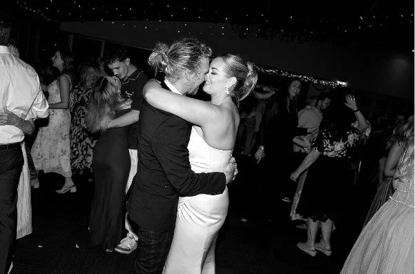 Two women dancing closely and embracing at a party, surrounded by other guests dancing and mingling.