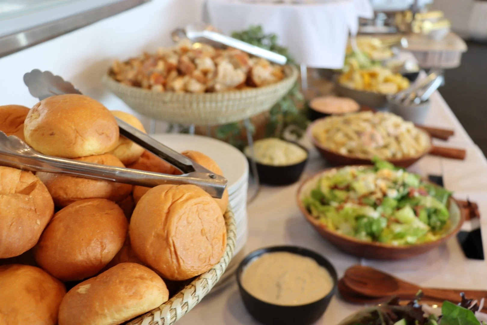 A bread basket with dinner rolls, and a buffet spread with various salads, pasta, and other dishes in bowls and platters on a long table.