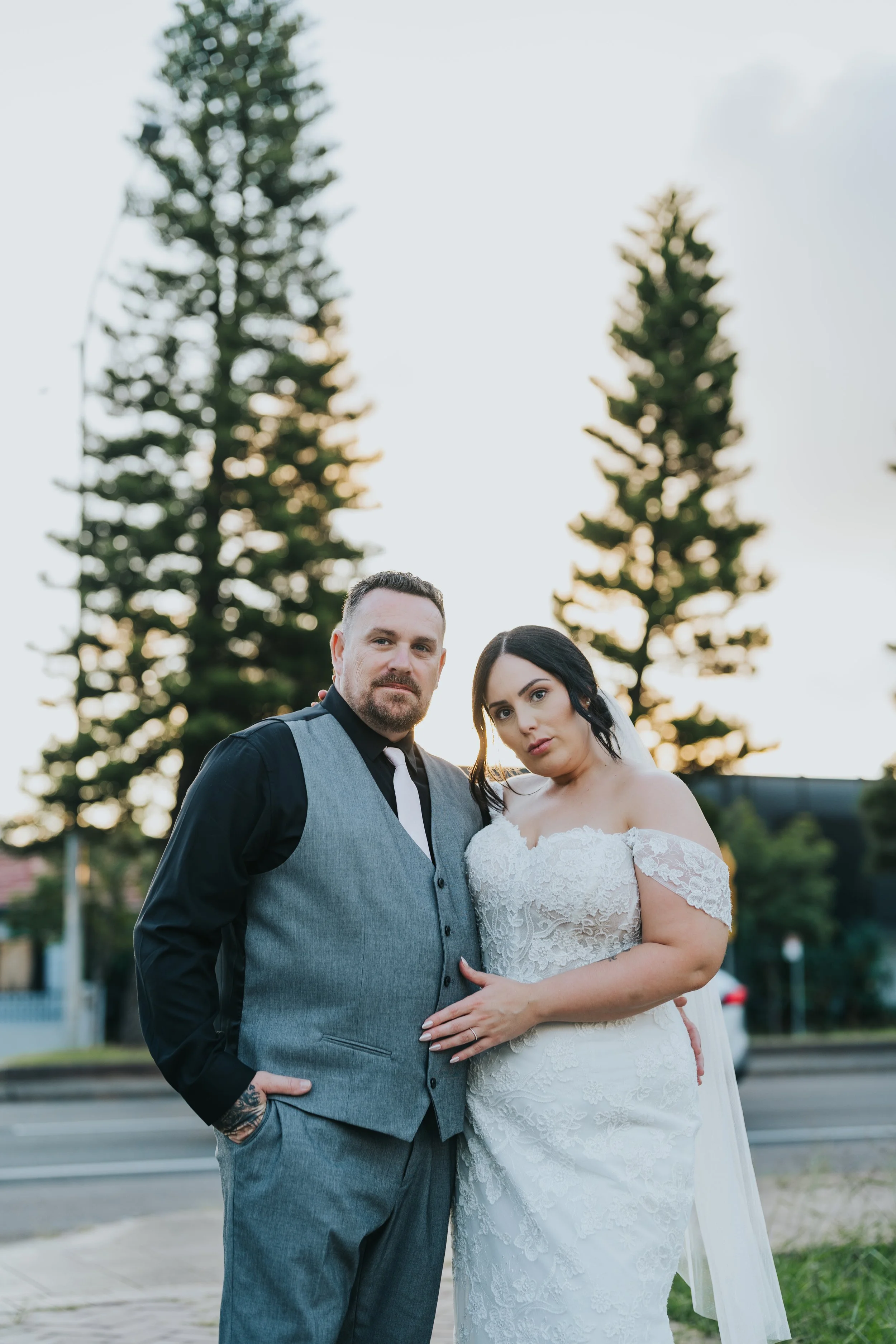 A bride and groom standing outdoors during sunset, with tall trees in the background, dressed in wedding attire.