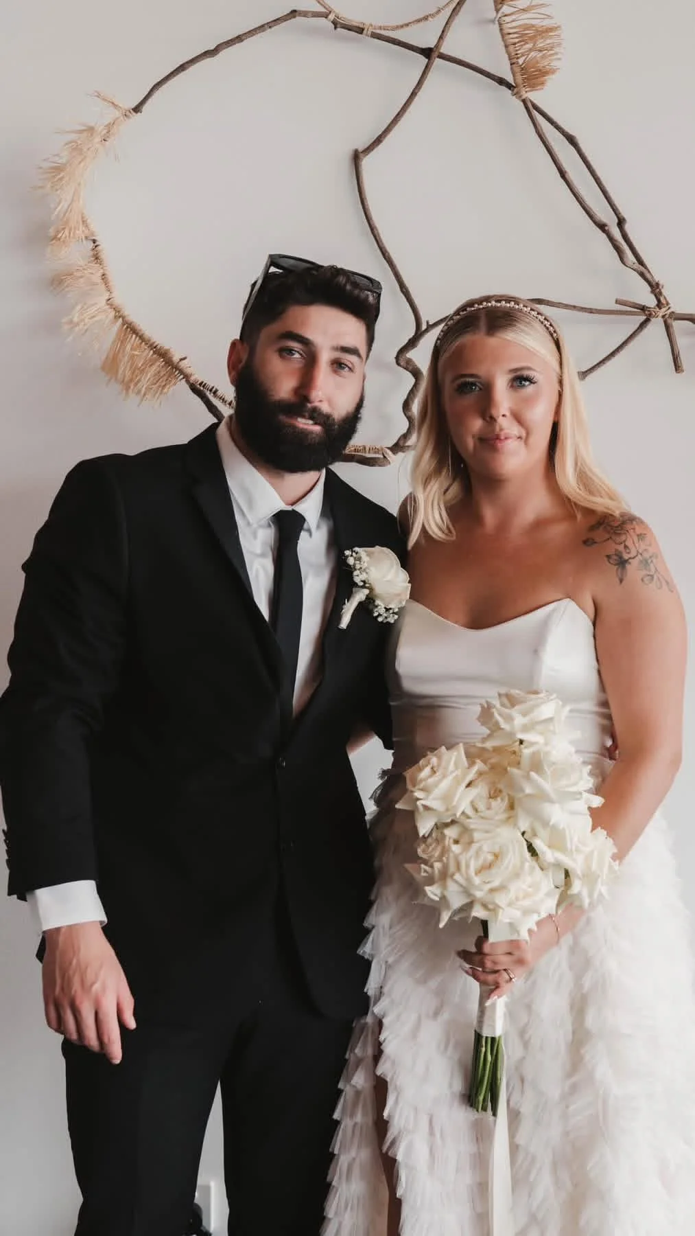 A bride and groom pose for a wedding photo indoors, with a decorative branch heart-shaped wall hanging in the background. The bride is holding a large bouquet of white roses, and the groom is wearing a black suit with a white shirt and black tie.