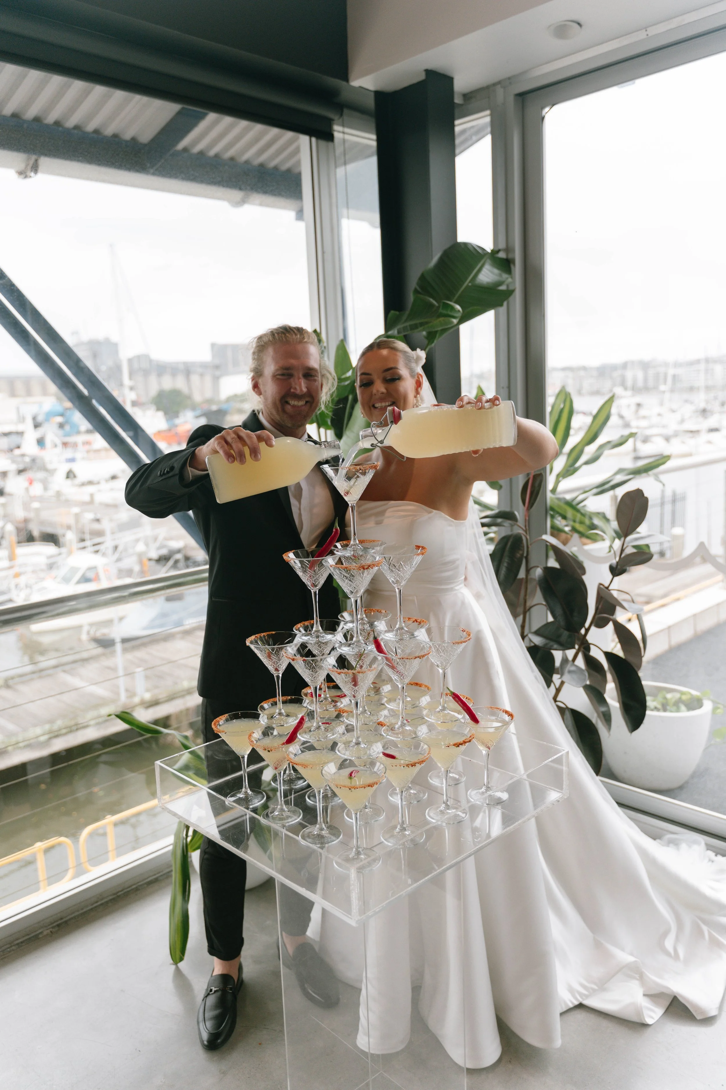 A newlywed couple is pouring yellow cocktail drinks into a pyramid of martini glasses during their wedding celebration. The bride is wearing a white wedding dress, and the groom is in a black suit. They are standing near large windows overlooking a m