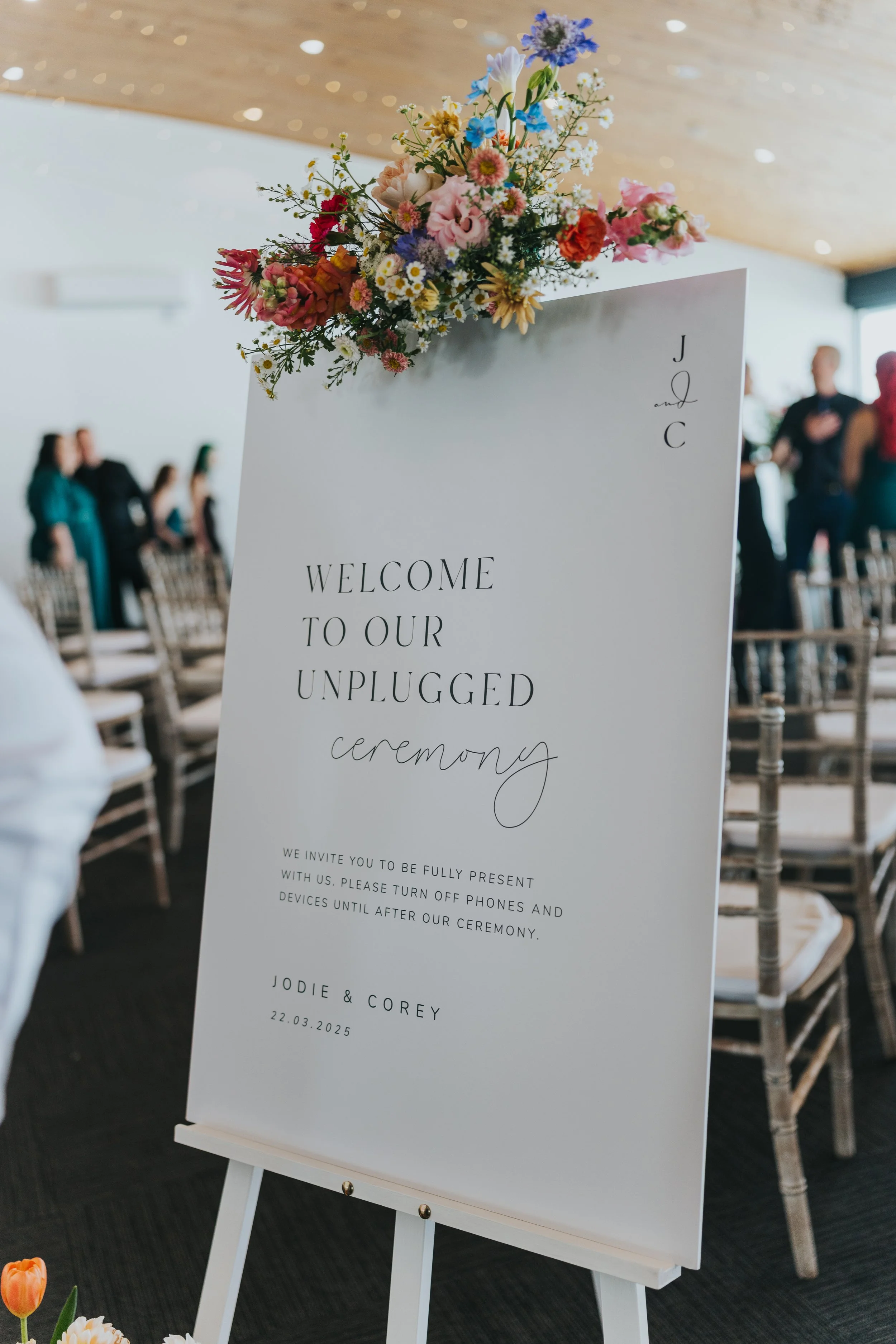Wedding welcome sign with floral arrangement on top, featuring pink, purple, blue, and white flowers. The sign invites guests to a ceremony on March 22, 2025, with instructions to turn off phones. Guests are shown in the background standing near chairs.