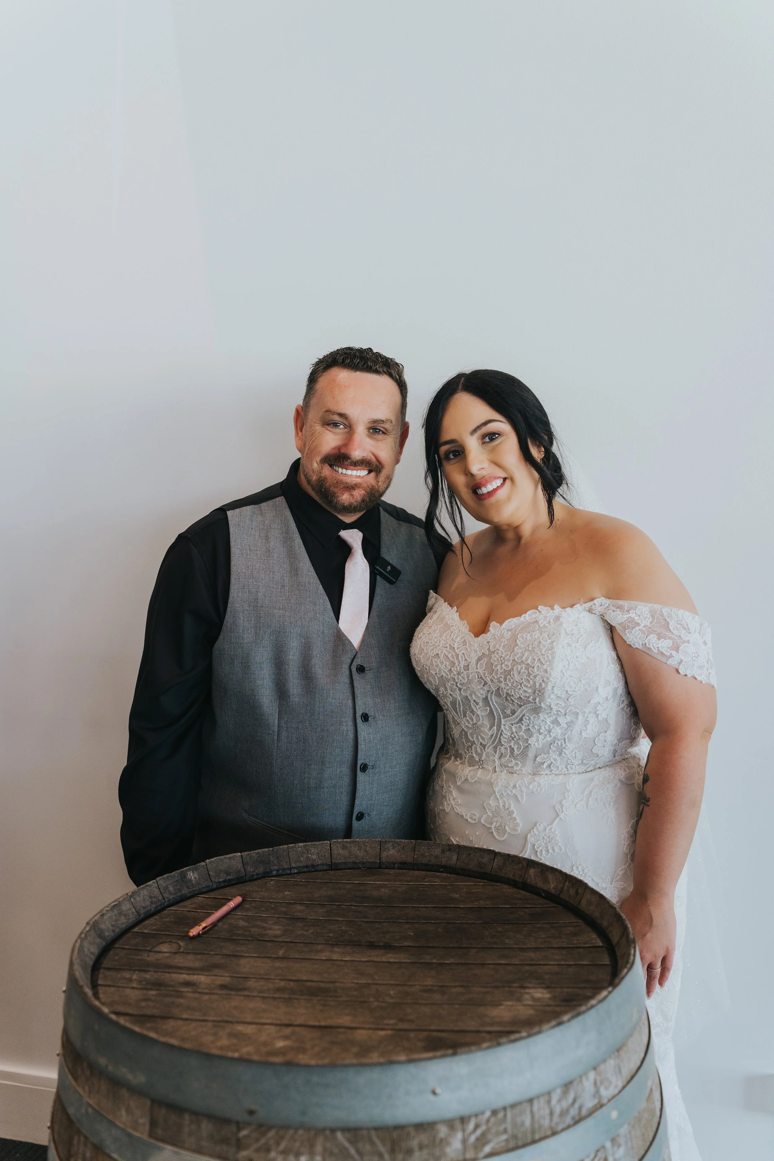 A smiling man in a grey vest and black shirt, and a smiling woman in a white lace wedding dress, standing together behind a wooden barrel, indoors with a plain white background.