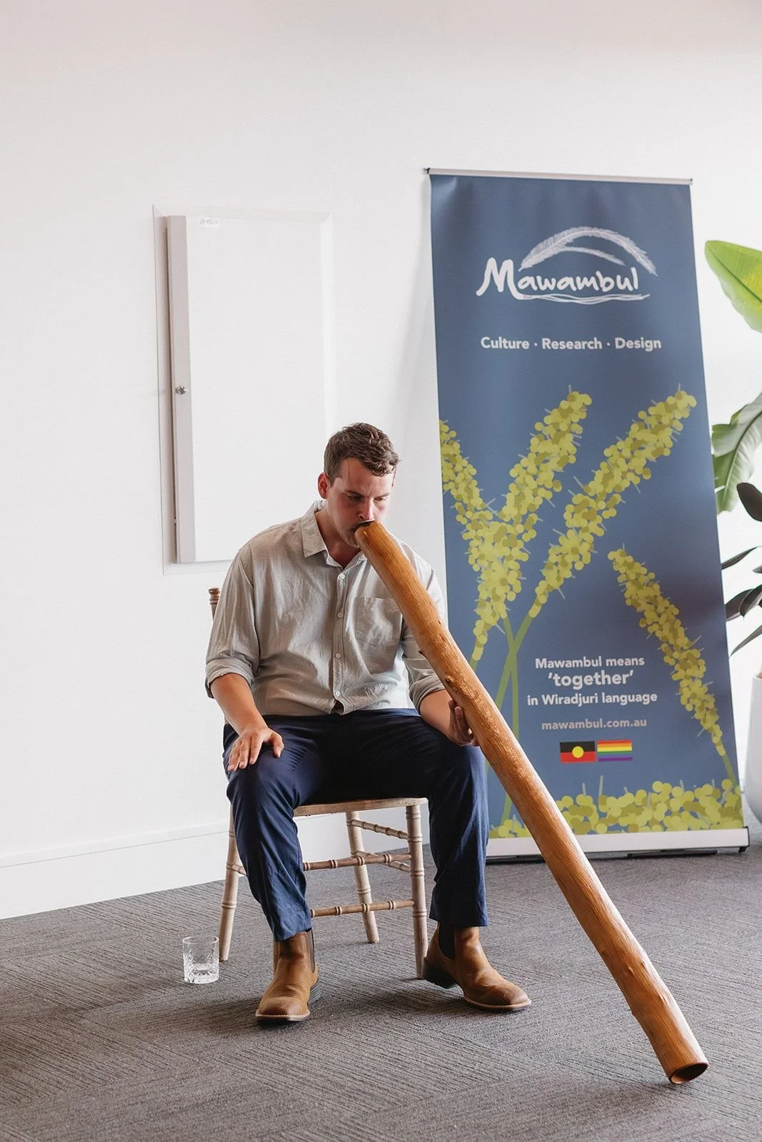 A young man sitting on a wooden chair, playing a long wooden didgeridoo, in front of a blue banner with yellow plant graphics and text about Mawambul culture and language.