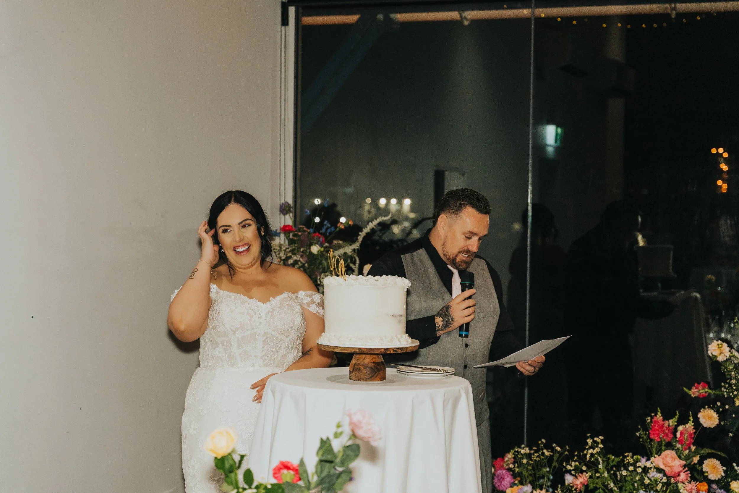A bride in a white lace wedding dress stands next to a man in a gray vest, who is reading from a paper and holding a microphone. They are at a wedding reception with a white cake on a round table, decorated with flowers and greenery.