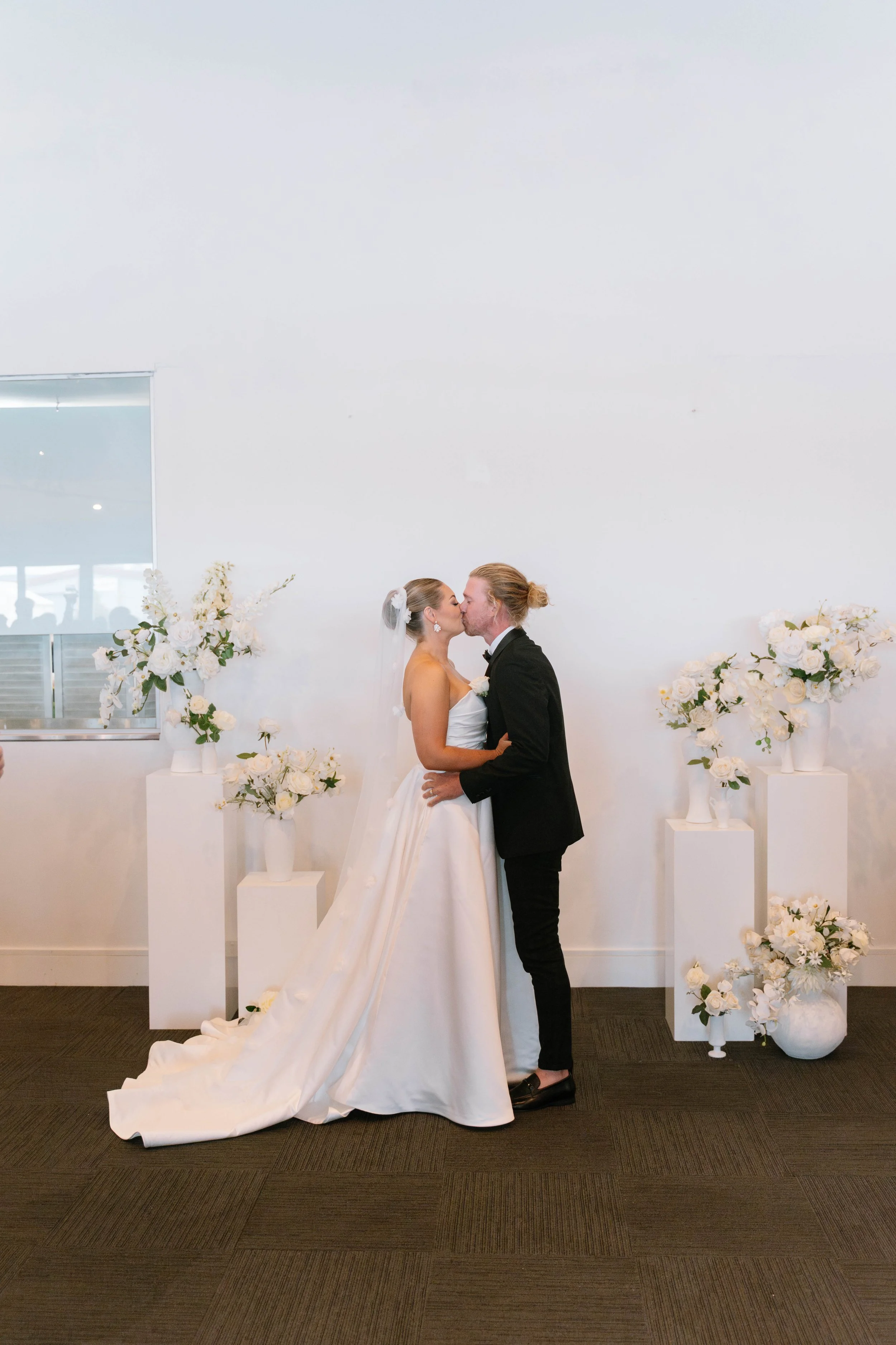 A bride and groom share a kiss at their wedding, surrounded by white floral arrangements.
