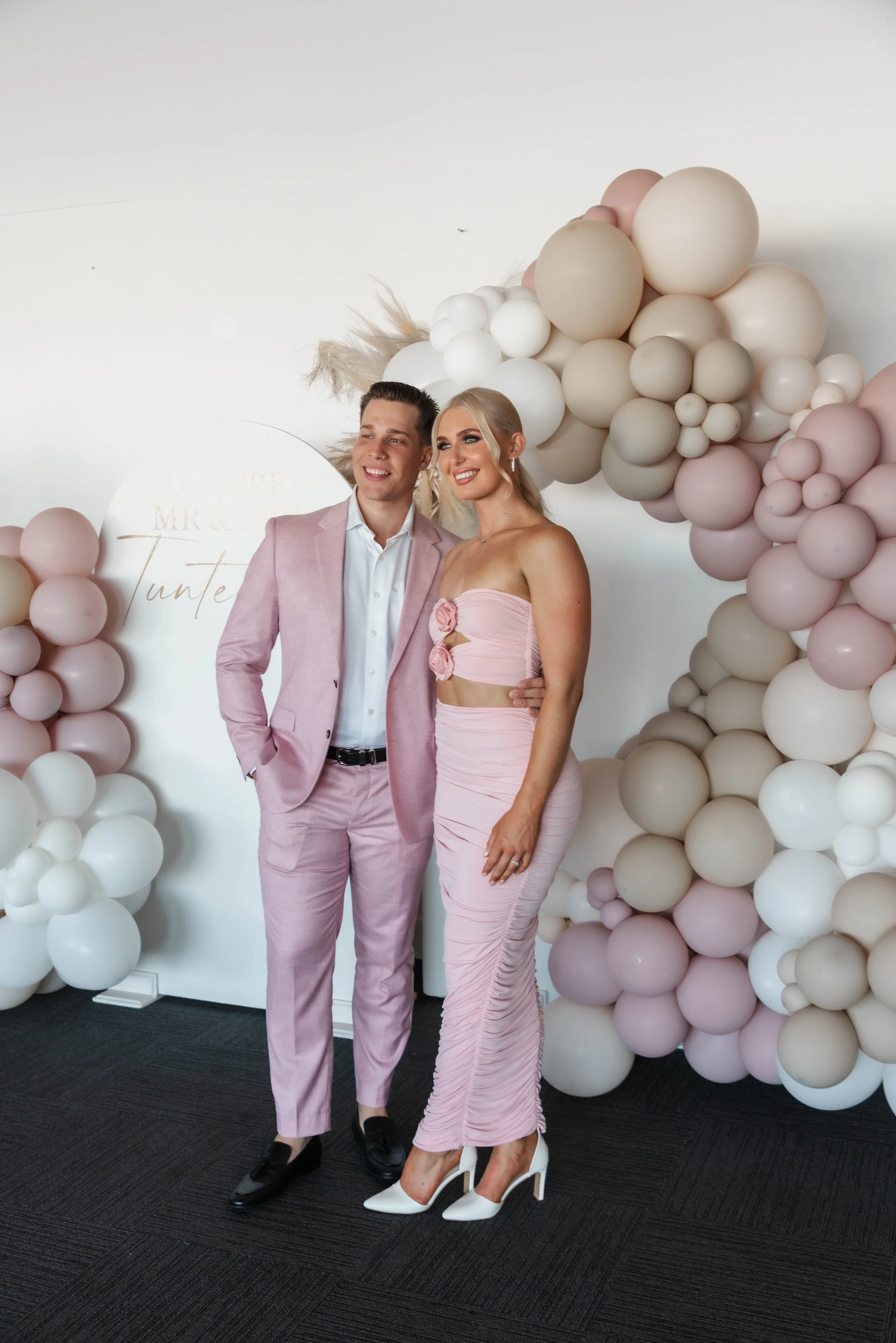A couple dressed in pink formal wear stands together in front of a balloon arch at a celebration, smiling.