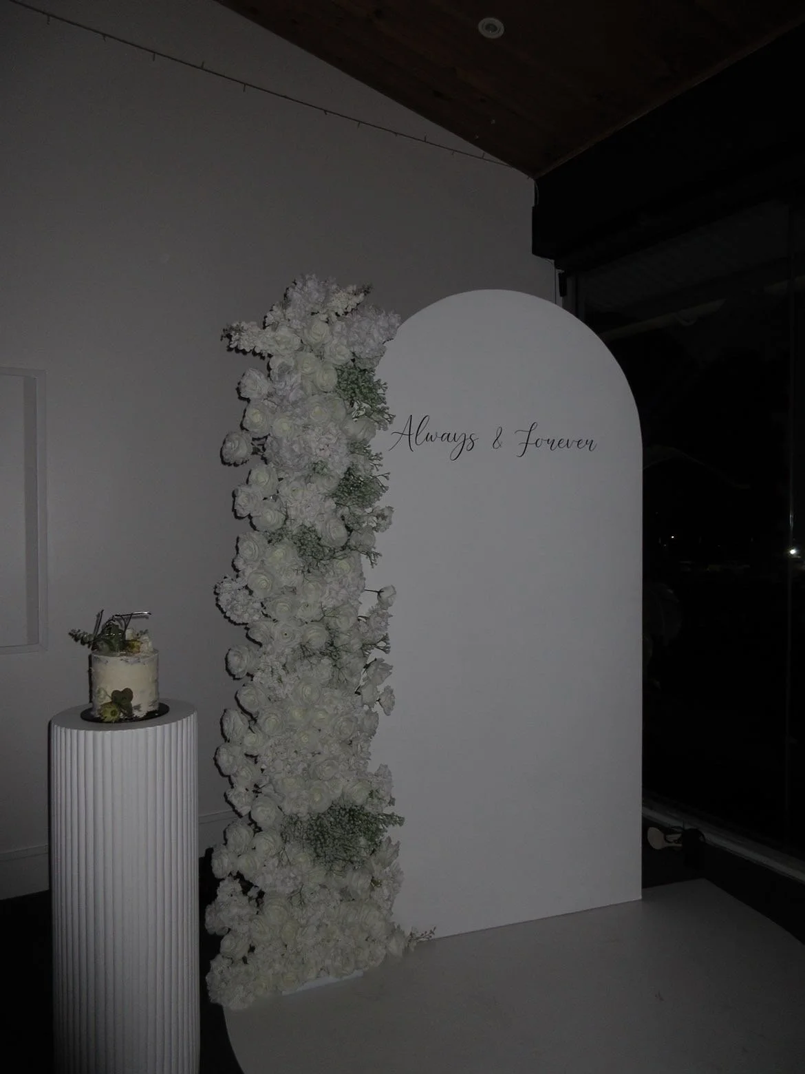 White floral arch with the inscription "Always & Forever" and a small white cake decorated with flowers sitting on a white pedestal.