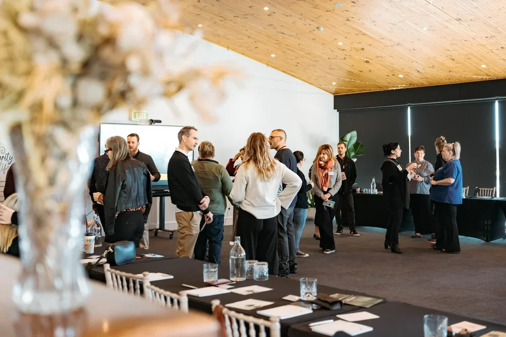 A group of people socializing indoors at a professional event or conference, with some people standing in small groups and others near a registration or check-in table.